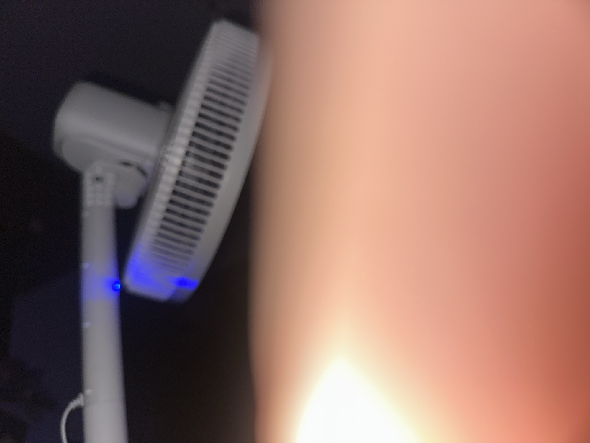 The image displays a close-up, out-of-focus view of a white pedestal fan. The fan's head, with its protective grille, is visible on the left side, angled upwards. A blue light is illuminated on the fan's base, possibly indicating it is powered on or in standby mode. The fan's vertical support pole is also visible, extending downwards. The majority of the image is dominated by a blurred, bright, peachy-orange area, which appears to be a hand or skin blocking a significant portion of the frame, suggesting the photo was taken by someone holding the camera. The background is dark and indistinct, offering no discernible environmental context. The image does not contain any elements that would allow for the identification of a specific location in Yerevan, Armenia, or any other city.