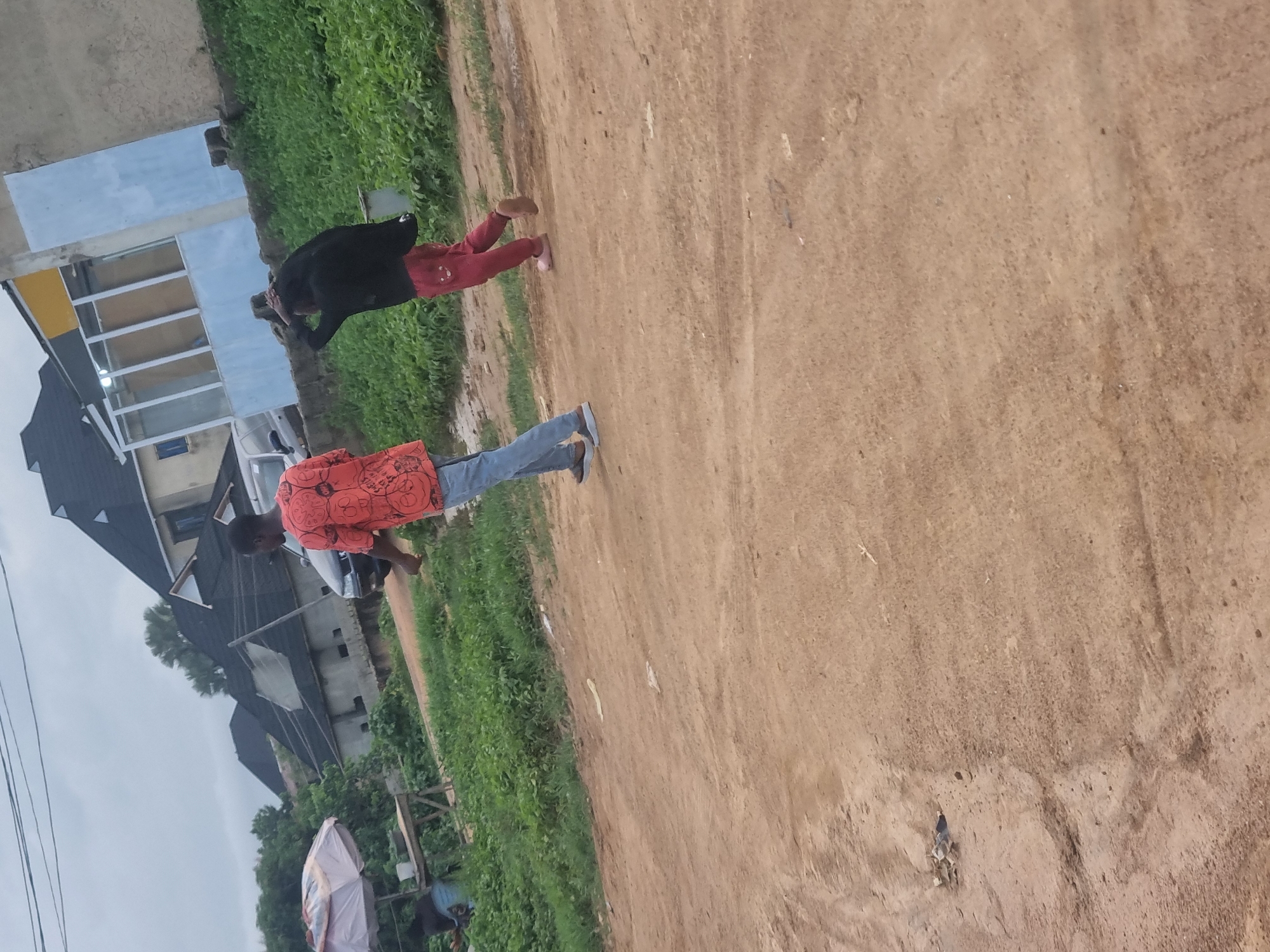 Two people are walking on a dirt path in Gwong, Nigeria. The person in the foreground is wearing an orange patterned shirt and blue jeans. The person in the background is wearing a black top and red pants. Behind the people, there are green bushes and some buildings under construction. The sky is overcast, suggesting it is either early morning or late afternoon. The ground is dry and dusty, with tire tracks visible in some areas. There is a white umbrella visible in the lower left corner of the image.