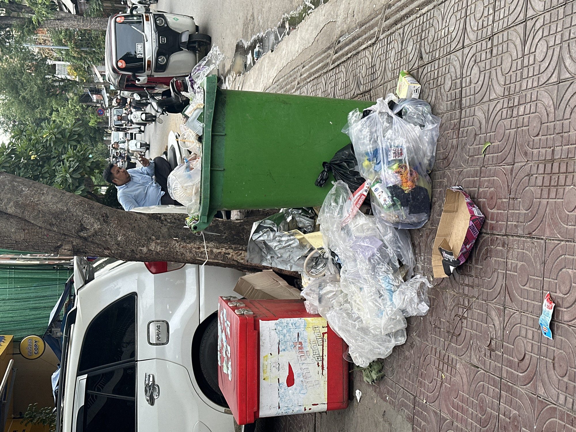 In Khan Daun Penh, Cambodia, a man in a light blue shirt and dark pants sits on the edge of an overflowing green garbage bin on a tiled sidewalk. He is looking down at his phone. To his left, a white SUV is parked. In front of the garbage bin, there is a pile of plastic bags filled with trash, along with a red cooler box. The street behind the man is busy with traffic, including tuk-tuks and motorcycles. There are trees and buildings lining the street.