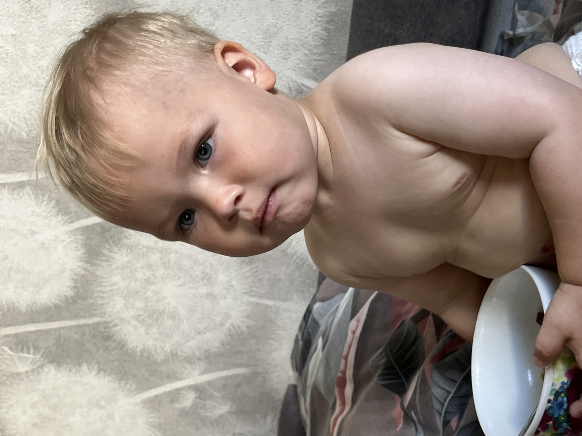 A baby boy with blond hair and blue eyes is sitting down, holding a bowl of food. He is looking to his right, with a slightly grumpy expression. The background appears to be a wall with a dandelion pattern. The baby is wearing a diaper and is shirtless. The image was taken in Chaikovsky, Russia.