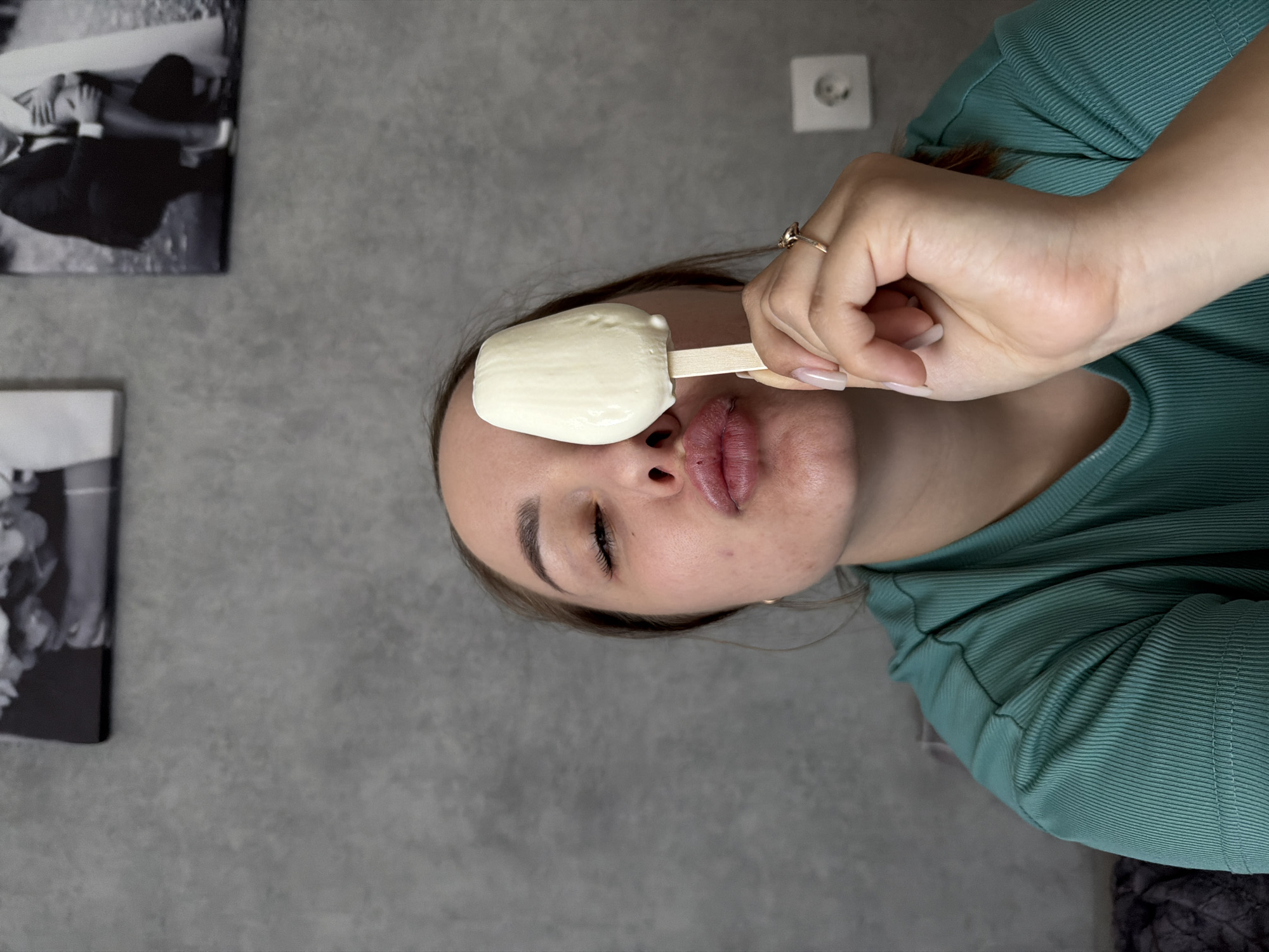 The image shows a woman holding a white ice cream bar on a wooden stick in front of her eye. She is wearing a green ribbed shirt and a gold ring on her finger. The background is a gray wall with two black and white framed photographs and an electrical outlet. The location is in Chaikovsky, Russia.