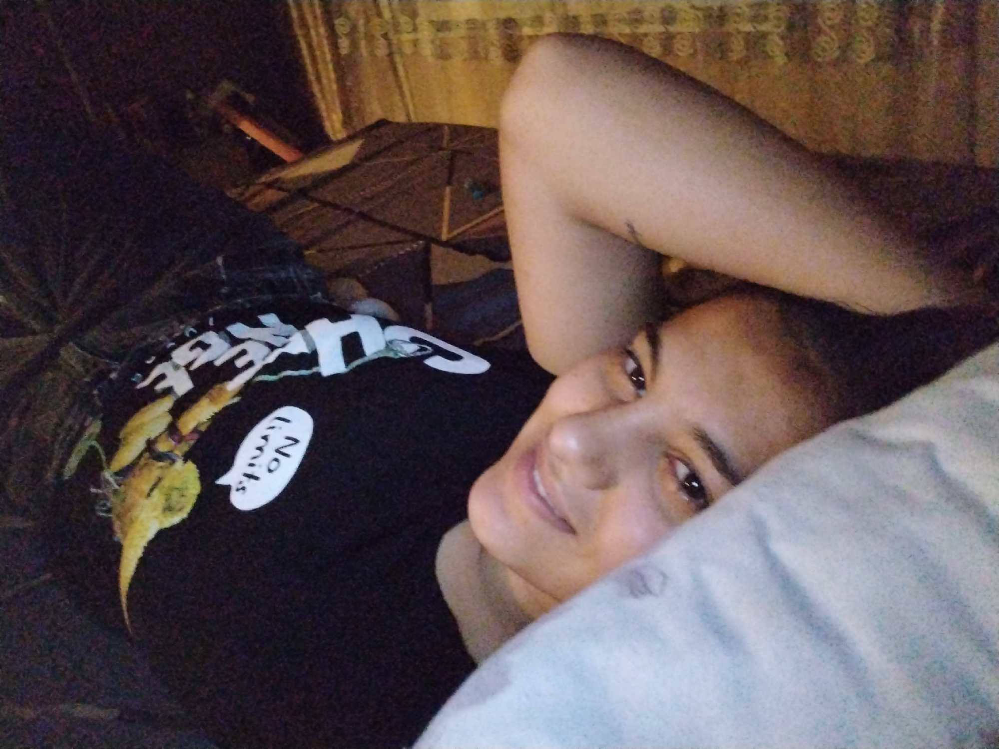 The image features a woman lying down indoors, likely in a bedroom setting, looking towards the camera. She is positioned on her side with her head resting on a light-colored pillow, partially covering her face. Her right arm is bent with her hand placed behind her head. She has dark hair, dark eyes, and a gentle, relaxed expression with a faint smile.
She is wearing a dark, presumably black, t-shirt. The t-shirt has a design featuring an abstract yellow, elongated figure or creature. Next to this design, a prominent white speech bubble contains the clear text "No limits." Other white text is partially visible on the t-shirt, but it is too obscured to be fully deciphered.
The background is dimly lit, suggesting either nighttime, early morning, or a darkened room. Behind the woman, a textured brown fabric, possibly a blanket or bedspread, is visible. The overall ambiance is tranquil and intimate.
Unknown, Unknown