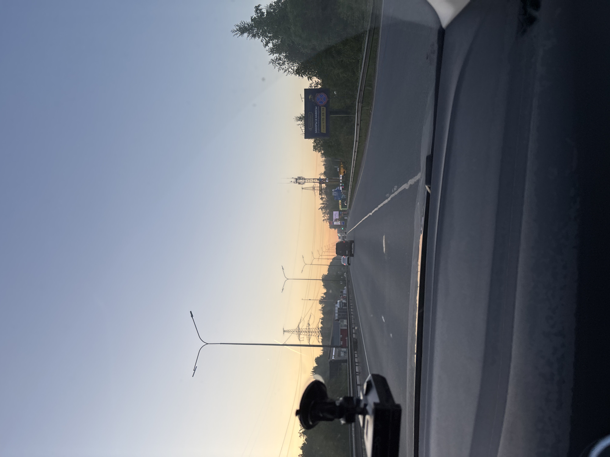 The image captures a view from inside a moving vehicle on a multi-lane highway, looking forward and slightly right. The perspective is from the driver's or front passenger's seat, with part of the dashboard and windshield visible on the right and bottom edges, along with what appears to be a dashcam mount in the lower center.
The sky exhibits a clear, pre-dawn or post-dusk glow, transitioning from light blue at the top to a warm orange-yellow closer to the horizon, indicating either sunrise or sunset. Trees and dense vegetation line both sides of the highway, separating it from the surrounding landscape. Numerous street lights with dual arms extend along the roadside, and several large power lines with their supporting towers are visible in the distance, running parallel to the road.
Vehicles can be seen on the highway, both ahead of the observer and in adjacent lanes, suggesting moderate traffic. One prominent billboard is visible on the right side of the road, displaying text in Russian: "ЗАБОРЫ И НАВЕСЫ" (ZABORY I NAVESY), which translates to "FENCES AND CANOPIES." Below this, a phone number, "8-900-650-70-10," is partially visible. The scene depicts a typical drive along a main road or highway in the early morning or late evening, consistent with an area like Zanevka, Russia, given the Cyrillic text.
