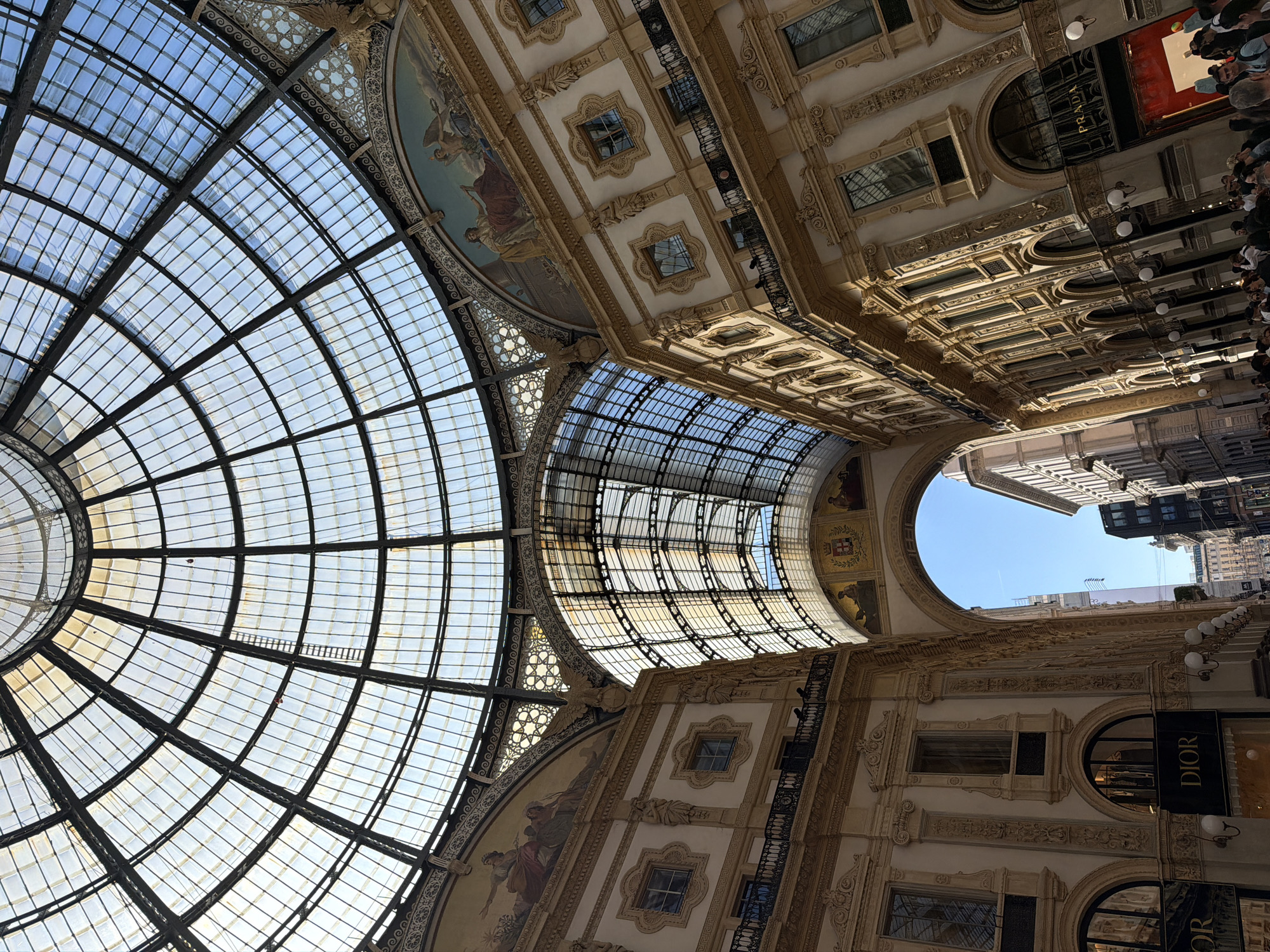 The image shows the interior of the Galleria Vittorio Emanuele II in Milan, Italy. The viewpoint is from below, looking up towards the glass and iron dome and the ornate surrounding architecture.
The glass dome features a grid pattern, allowing natural light to filter through. The architecture includes detailed plasterwork, paintings, and window openings, with the luxury brand stores like Prada and Dior visible along the walls. A crowd of people can be seen along the edge of the frame, suggesting a busy commercial area. At the end of the gallery, a portion of the open sky and surrounding buildings are visible.
