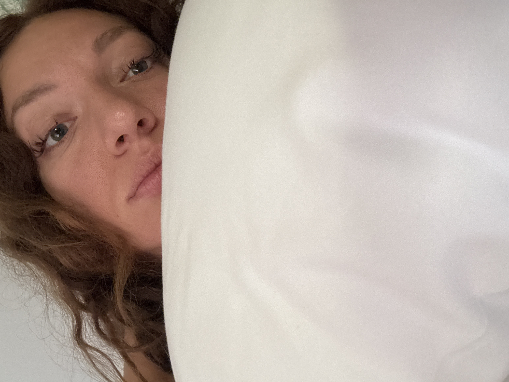 A woman's face is partially visible, with her eyes, nose, and a portion of her mouth showing. She has curly brown hair. A large white object obscures the right side of the frame, possibly a pillow or blanket. The environment is nondescript, giving no indication of the image being taken in Đà Nẵng, Vietnam.