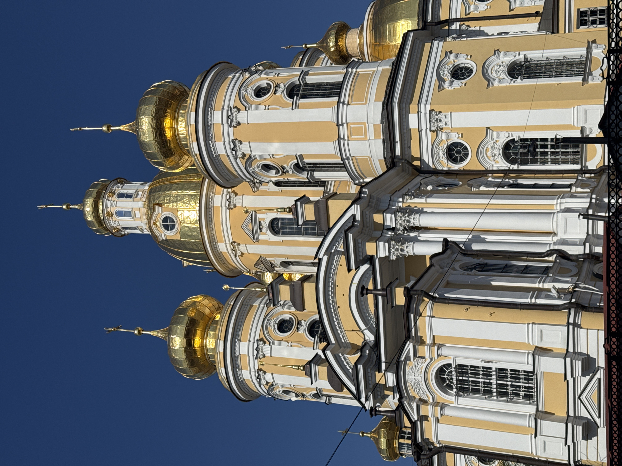 The image shows the gold domes and ornate architecture of a building, likely a church or cathedral, set against a clear blue sky. The building is decorated with yellow and white facades, with detailed architectural elements, including windows, columns, and moldings. The gold domes are particularly prominent, catching the sunlight and reflecting a bright, metallic sheen. The image is shot from a low angle, emphasizing the height and grandeur of the structure. Based on the architectural style and gold domes, it is likely located in Saint Petersburg, Russia.