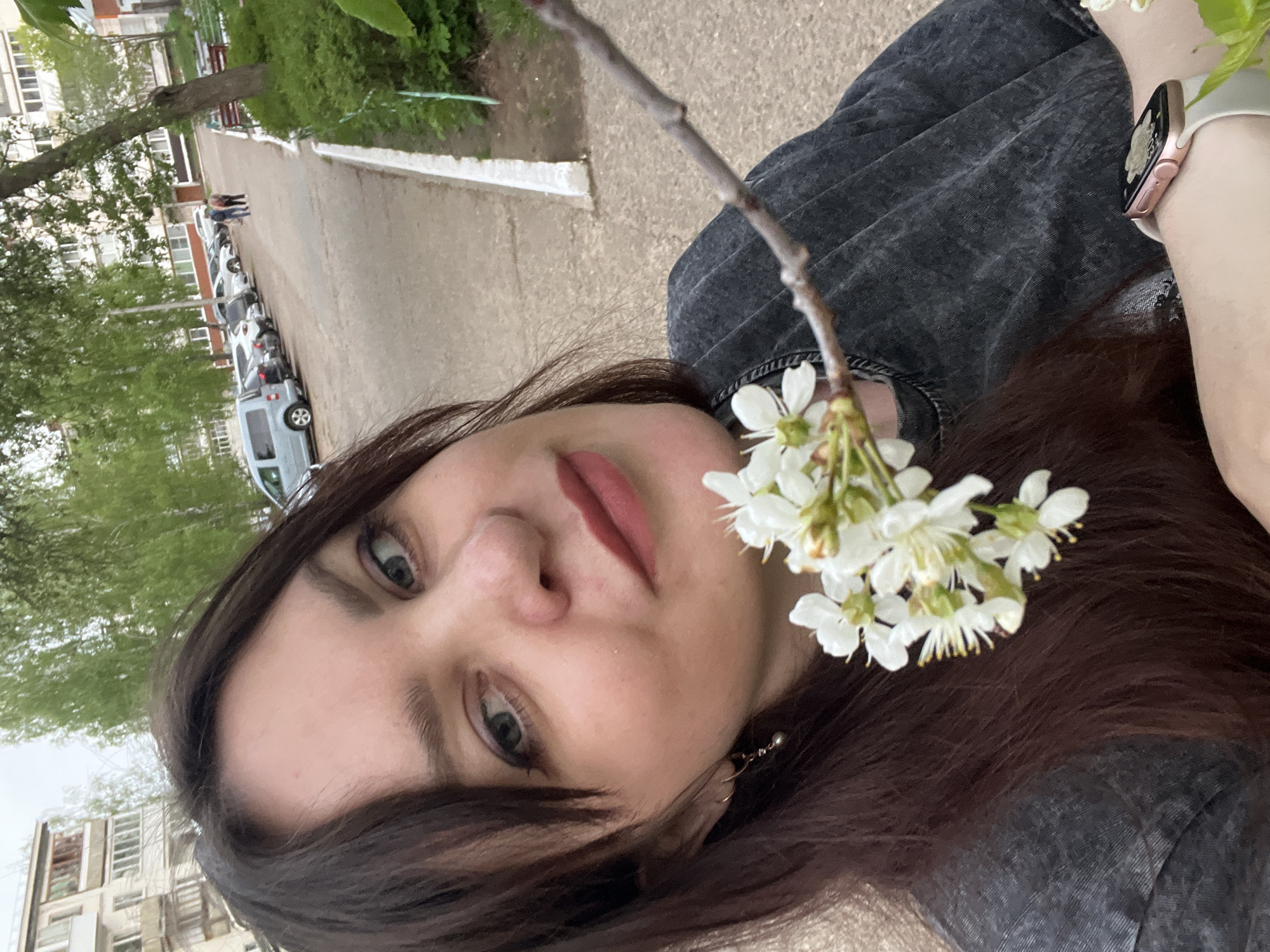 A selfie depicts a woman with dark hair and light skin, wearing a black denim jacket and an Apple Watch with a white band. She is holding a flowering tree branch in front of her. The background shows a street in Noviy, Russia with buildings, parked cars, and trees visible. Two people are walking in the distance.