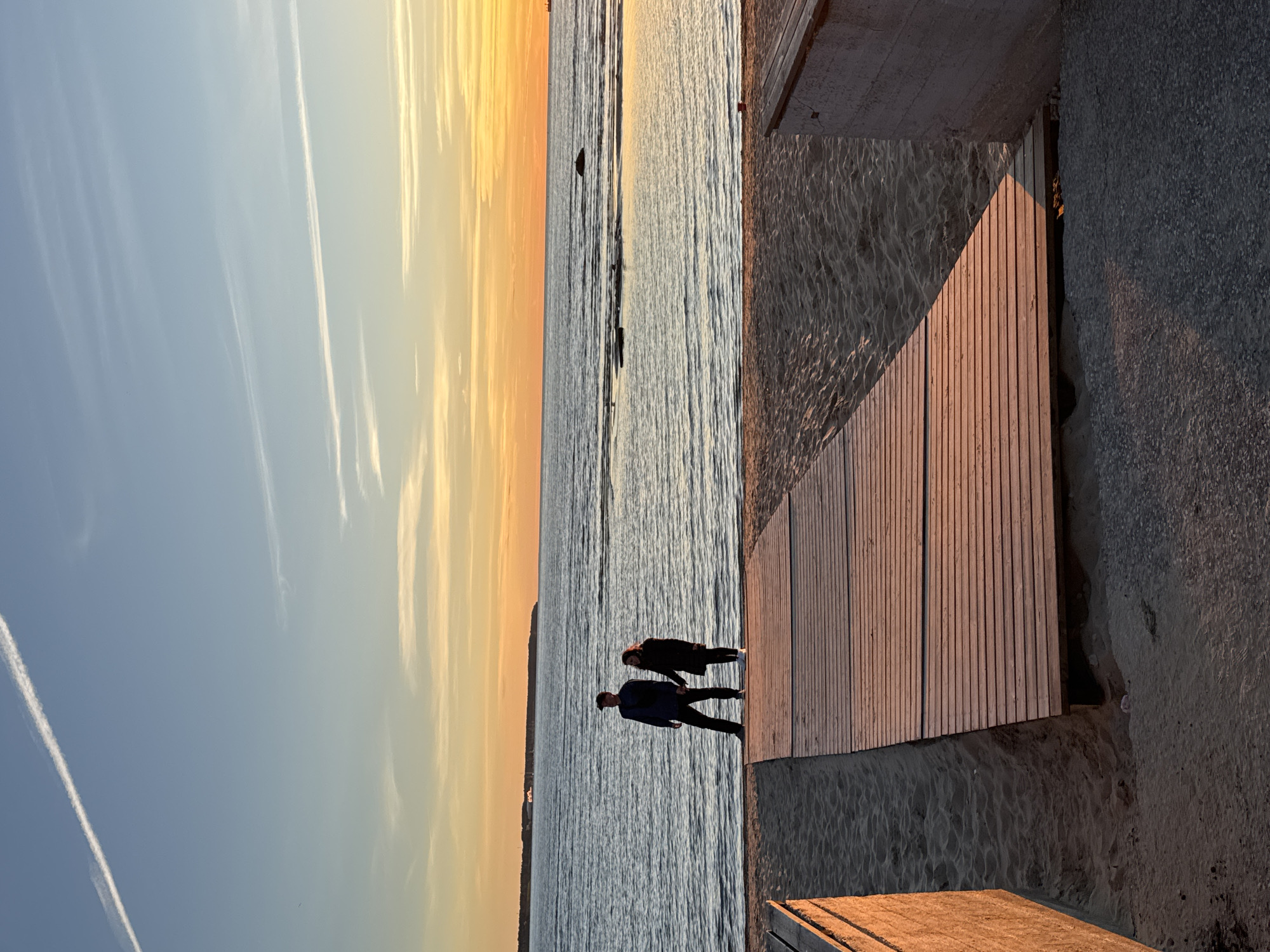 Two individuals, a male and a female, stand on a wooden boardwalk at a sandy beach, facing the sea during sunset. The male, wearing a dark jacket and pants, stands slightly to the left and ahead of the female, who is dressed in a dark coat and pants. They appear to be holding hands. The boardwalk, constructed from light-colored wooden planks, extends from the foreground towards the sea. The beach is sandy, with some debris visible near the boardwalk's edge. The sea is calm, with gentle waves. In the background, the sky displays a gradient of colors from blue at the top to orange and yellow near the horizon, indicating sunset. A distant coastline with some structures is visible, and a ship is present on the horizon. The location is identified as Tallinn, Estonia. The lighting suggests it is late afternoon or early evening.