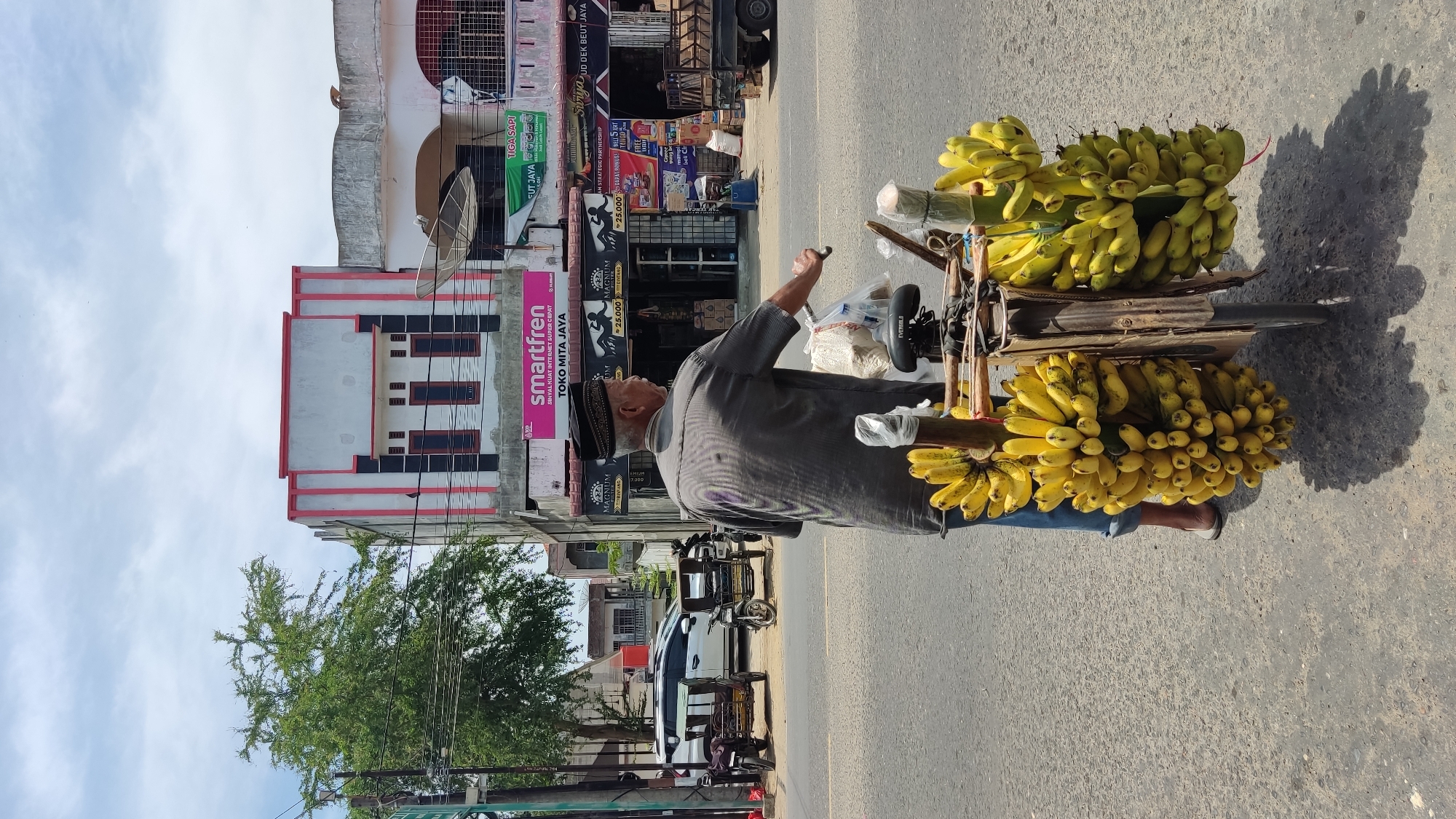 An elderly man is pedaling a heavily loaded bicycle on a street in what appears to be Lhokseumawe, Indonesia. He is wearing a striped gray shirt, blue jeans, a taqiyah, and white shoes. The bicycle is loaded with multiple bunches of ripe yellow bananas. The man is pedaling along a paved street in front of several shops. Prominent store signs in the background include those for "smartfren" and "Surya UD DEK BEUT JAYA". Several parked motorcycles and a white car can be seen further down the street. The sky is partly cloudy.