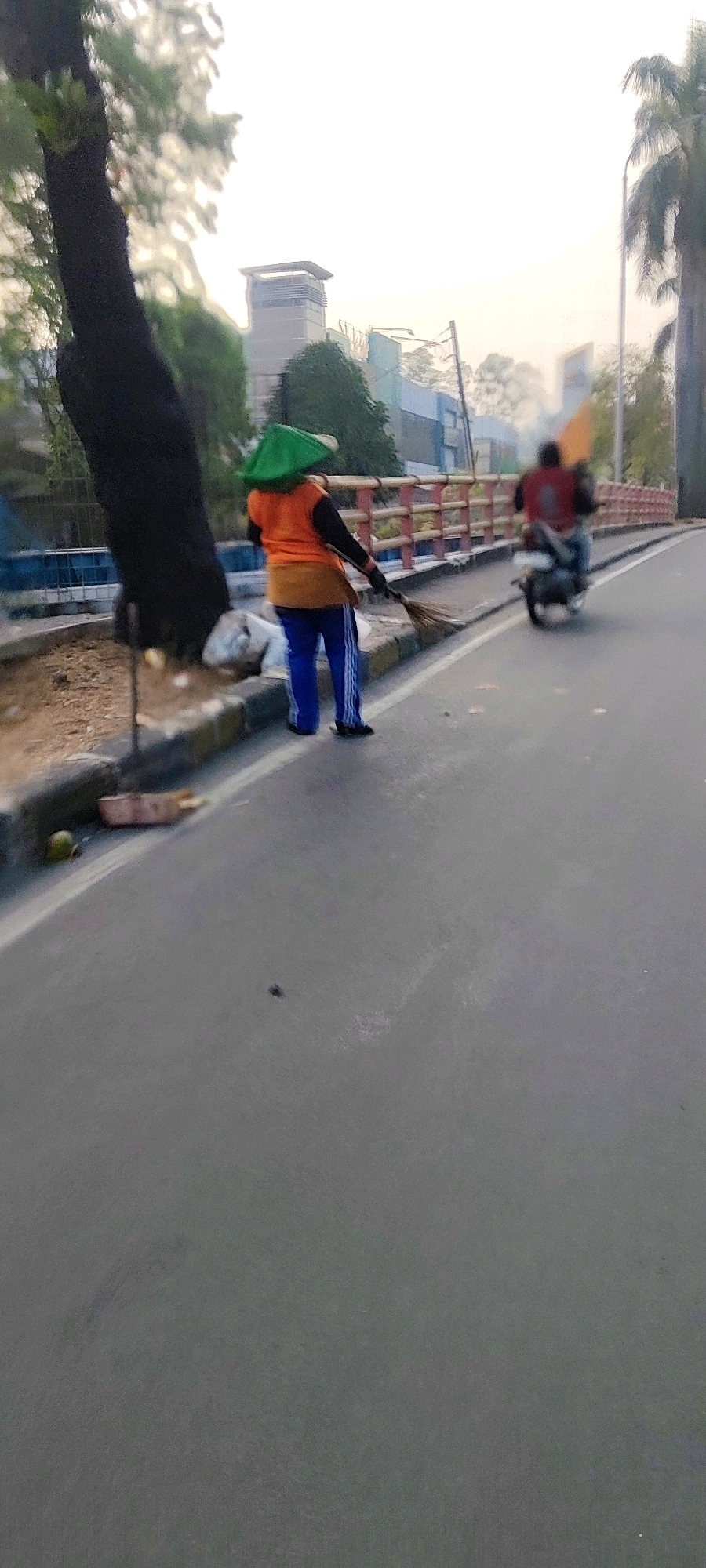 In Mekarmukti, Indonesia, a street cleaner wearing an orange vest, brown apron, and blue pants with white stripes sweeps the side of a road. They also wear a green conical hat. The person is standing next to a tree and a pile of trash bags. A motorcycle with a person on it is driving away on the road. The scene is set against a backdrop of buildings and palm trees under a bright sky.