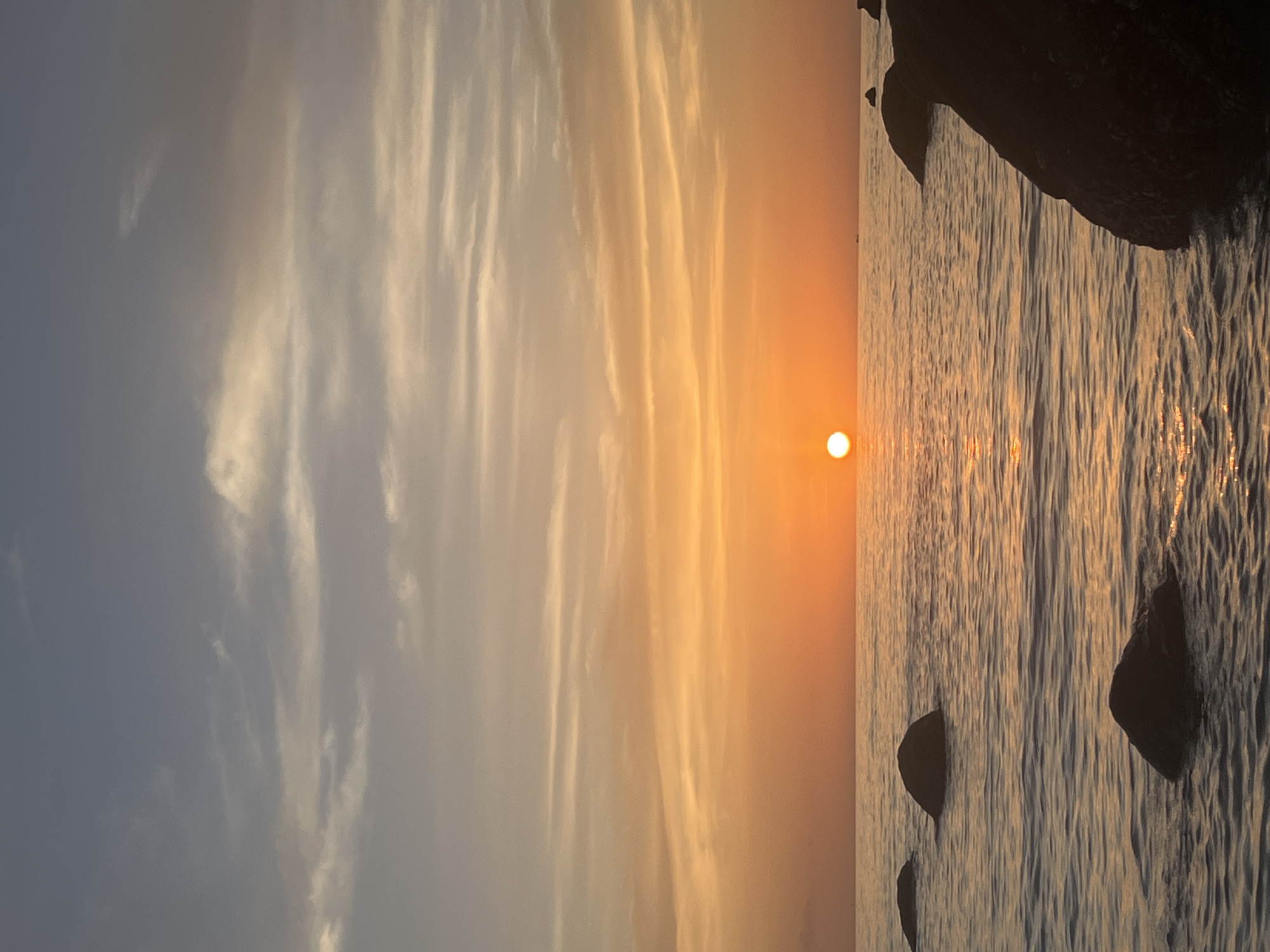 The image captures a serene sunset over the ocean in Ko Pha-ngan District, Thailand. The sun, a bright orange orb, is positioned low on the horizon, casting a warm glow across the sky and water. Wispy clouds are scattered across the upper portion of the frame, their colors ranging from pale blue to soft orange. The sea is calm, with gentle ripples reflecting the sunset's hues. Several large, dark rocks are visible, some partially submerged in the water, adding a naturalistic element to the seascape. The overall impression is one of tranquility and natural beauty, characteristic of a tropical sunset.