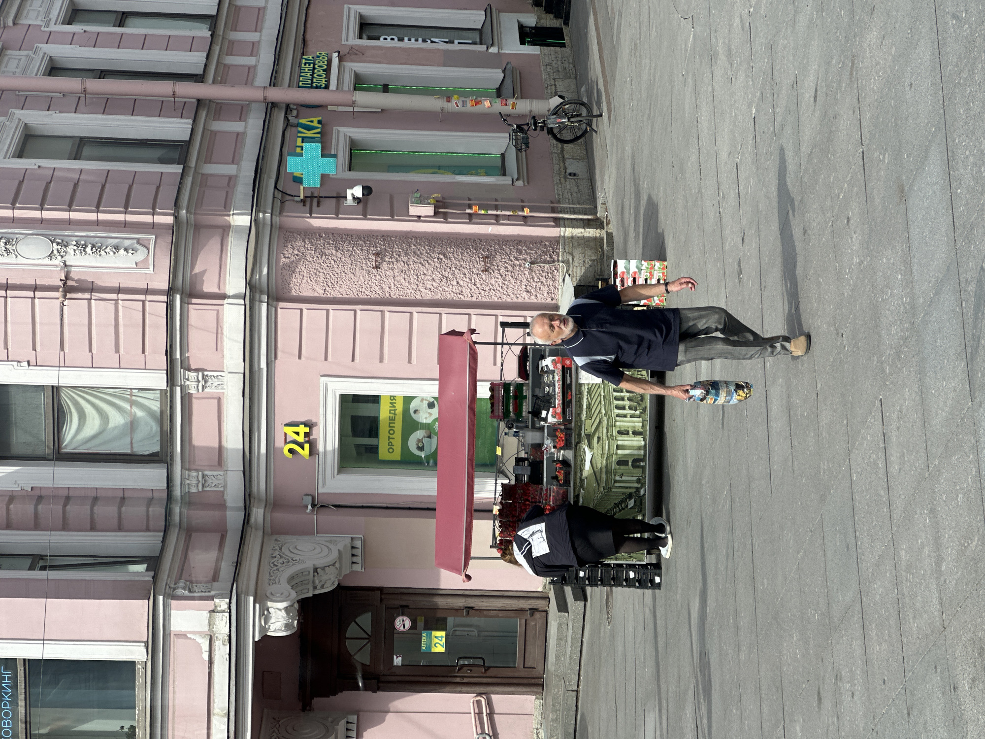 The image shows a street scene in Saint Petersburg, Russia. A pink building stands prominently on the left with a sign in Russian that includes the word "АПТЕКА" (pharmacy) and a green cross, indicating a pharmacy. The building also displays the number "24" in yellow. A person, possibly a vendor, stands with a display of what appear to be fruits or vegetables. Another person appears to be stocking or organizing goods near the storefront. On the right, a man stands holding an item, possibly a bottle. The environment includes a gray pavement and a bicycle parked near the building.