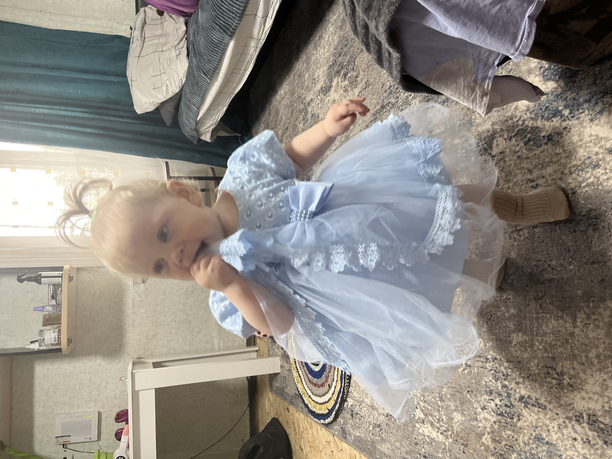 A young Caucasian toddler with blonde hair pulled into pigtails stands on a patterned rug in a room. She is wearing a light blue, lacy tutu dress with pearl embellishments and a large bow at the waist. Her face is turned towards the camera, and she is putting her hand to her mouth. The background shows a messy room with a bed in the upper left corner, covered with pillows and blankets in shades of grey, white, and purple. To the left of the bed, teal curtains hang near a window with light streaming through. Below the window, there is a shelf with various items, including bottles and what appear to be crafting supplies. A white table or shelf unit is visible in the lower right corner, and a small, round rug with concentric circles of color lies on the floor in front of the toddler. The overall impression is an indoor, casual setting. No specific time of day or weather conditions are evident, but the lighting suggests it might be daytime. The image appears to be taken in a residential environment in Фоки, Russia. There is no visible text in the image.