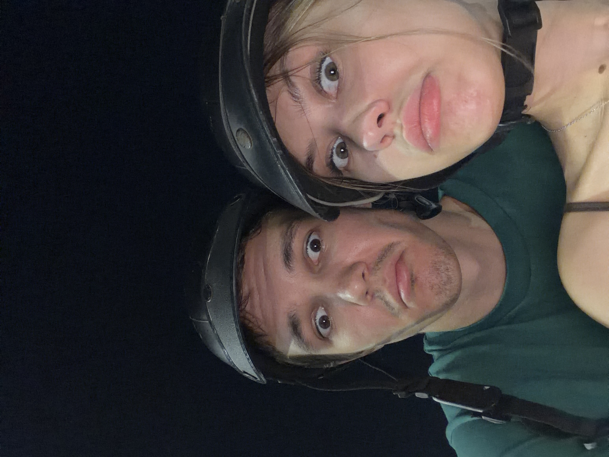 Two individuals, a male and a female, are captured in a close-up selfie, both wearing helmets and making humorous, surprised facial expressions with wide eyes and pouty lips. The background is entirely black, suggesting a nighttime setting or a dark environment. The man on the left wears a dark green t-shirt, and the woman on the right has a dark-colored strap visible across her chest, possibly from a top or bra. The image is of low resolution. No discernible details of Đà Nẵng, Vietnam, are present in the image.