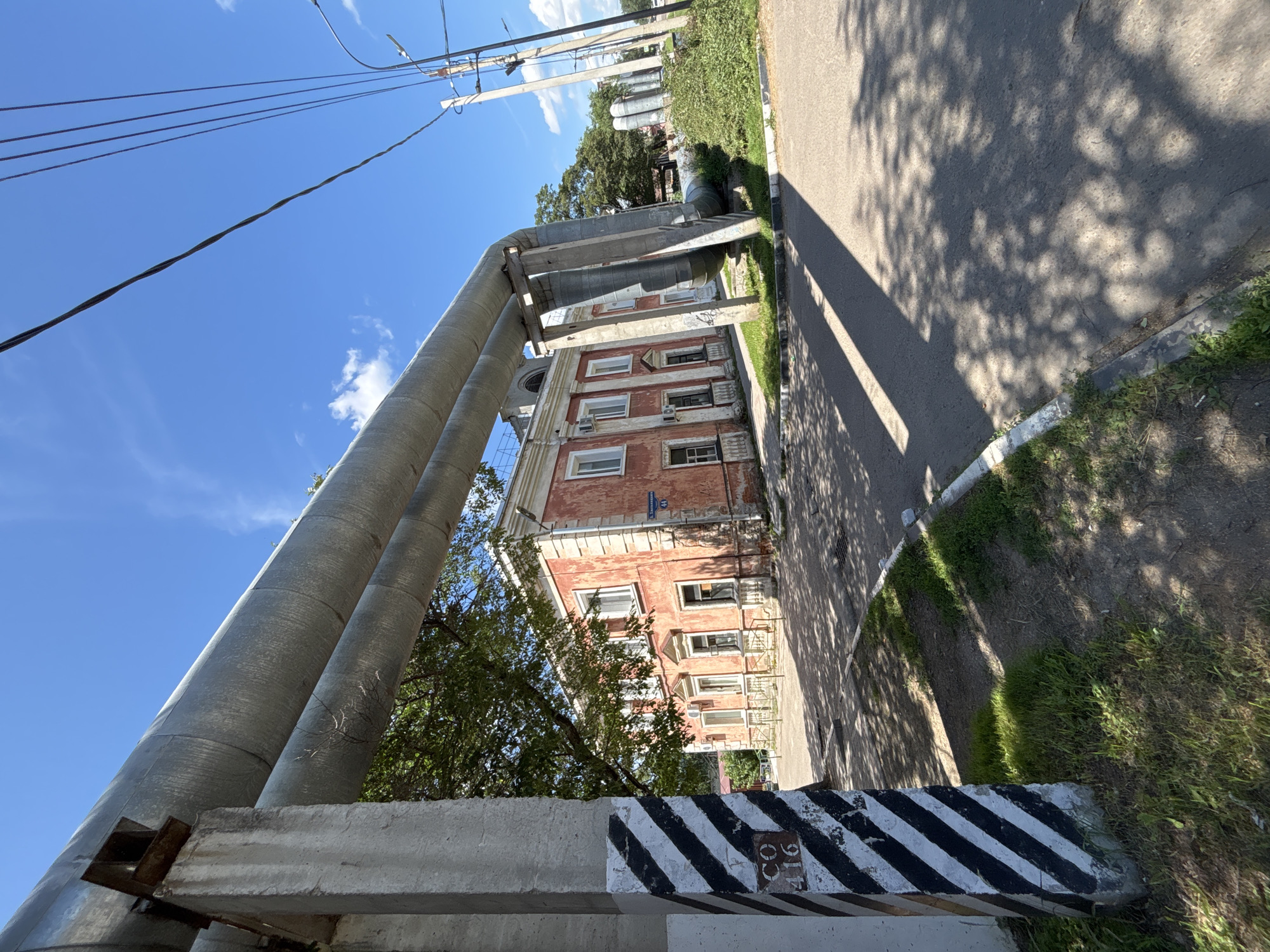 The image shows a street scene in Blagoveshchensk, Russia, featuring a large industrial pipe system running overhead. The pipes are silver and supported by concrete pillars. A two-story building with a reddish-pink facade and white trim is visible behind the pipes, with multiple windows and ornate architectural details. A street sign is affixed to the building, indicating "G. Shchetinkina" and the number "44". A concrete pillar in the foreground, painted with diagonal black and white stripes, has the inscription "CO 416" visible. Power lines are strung across the bright blue sky, which also has a few scattered clouds. The ground is asphalted, with some patches of grass and greenery. Shadows from trees and the pipes are cast across the road. The overall impression is an industrial or utility area within an urban environment.