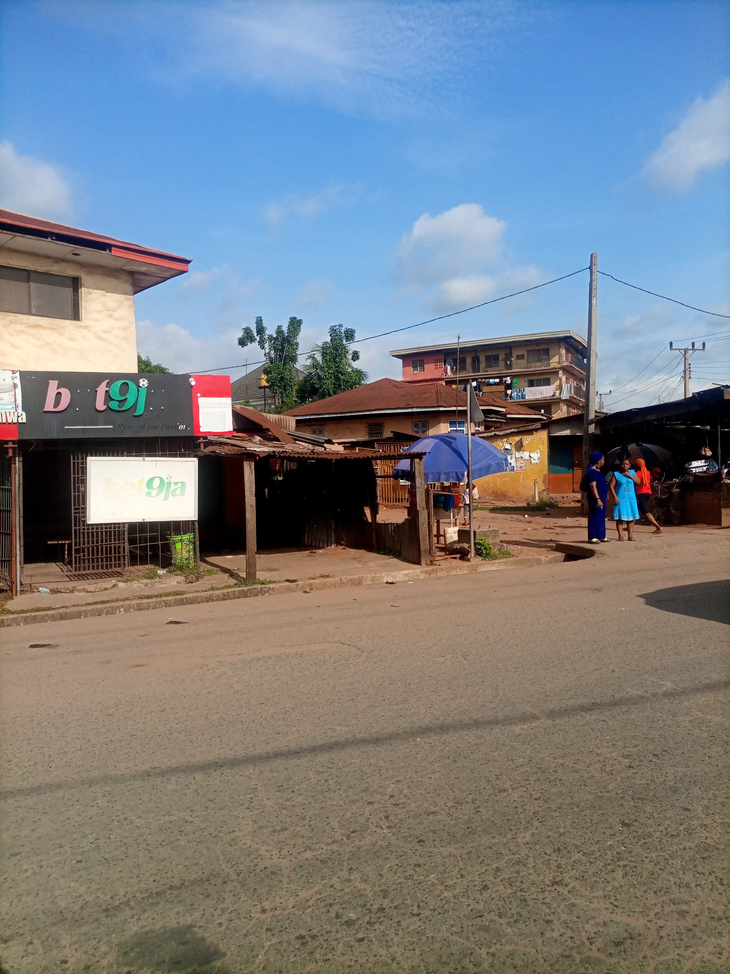 The image captures a street scene in Nkpor, Nigeria, under a bright, cloudy sky, suggesting it is daytime. The foreground shows a paved road with a worn surface, and a concrete curb to the left.
On the left side of the image, a building with a light-colored facade and a red trim is visible. Attached to this building is a dark sign with colorful text that reads "b t9j" and a smaller slogan below that says "Reward for Passion." Below this sign, a white board displays "bet9ja" in green. This area appears to be a commercial establishment, possibly a betting shop. A metal gate and some fencing are also present.
Further into the scene, several structures with corrugated iron roofs are lined up, indicating a market or informal commercial area. A blue umbrella shades a small stall. In the middle ground, three people are walking and standing on the dirt path beside the road. One person is wearing a bright orange shirt, another a blue dress, and the third a dark blue outfit. Their posture suggests casual interaction.
In the background, a multi-story building with balconies and laundry hanging out can be seen, along with utility poles and wires. The overall impression is of a busy, everyday street life in a Nigerian town.