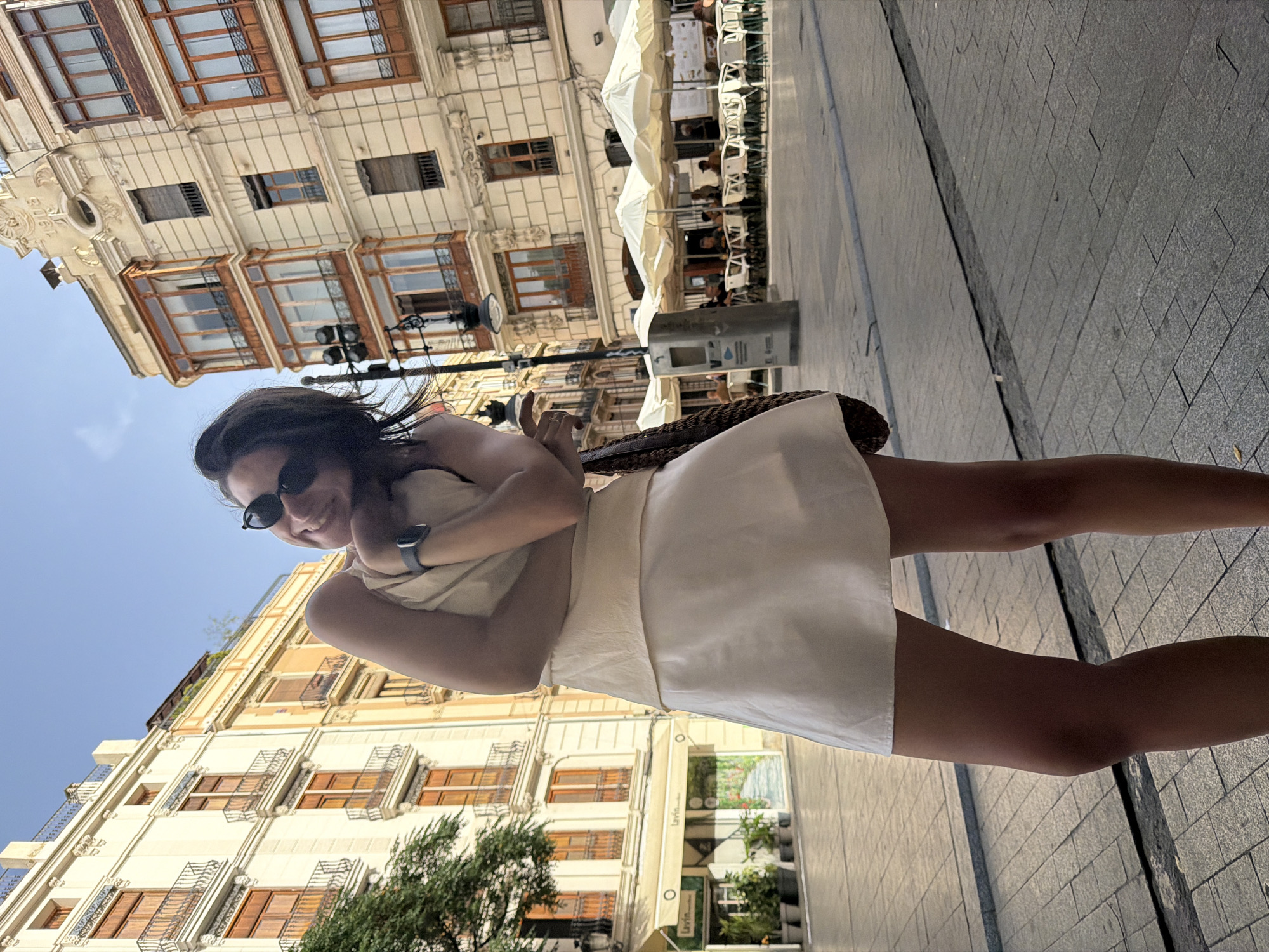 A young woman with dark hair and sunglasses smiles at the camera while standing on a cobblestone street in an urban setting. She is wearing a light-colored sleeveless top and a short skirt. Her right arm is bent, and she rests her chin on her hand. A woven bag is slung over her left shoulder. In the background, ornate buildings with balconies and large windows line the street. Outdoor seating with umbrellas and tables is visible in front of a cafe. The sky above is clear and blue.