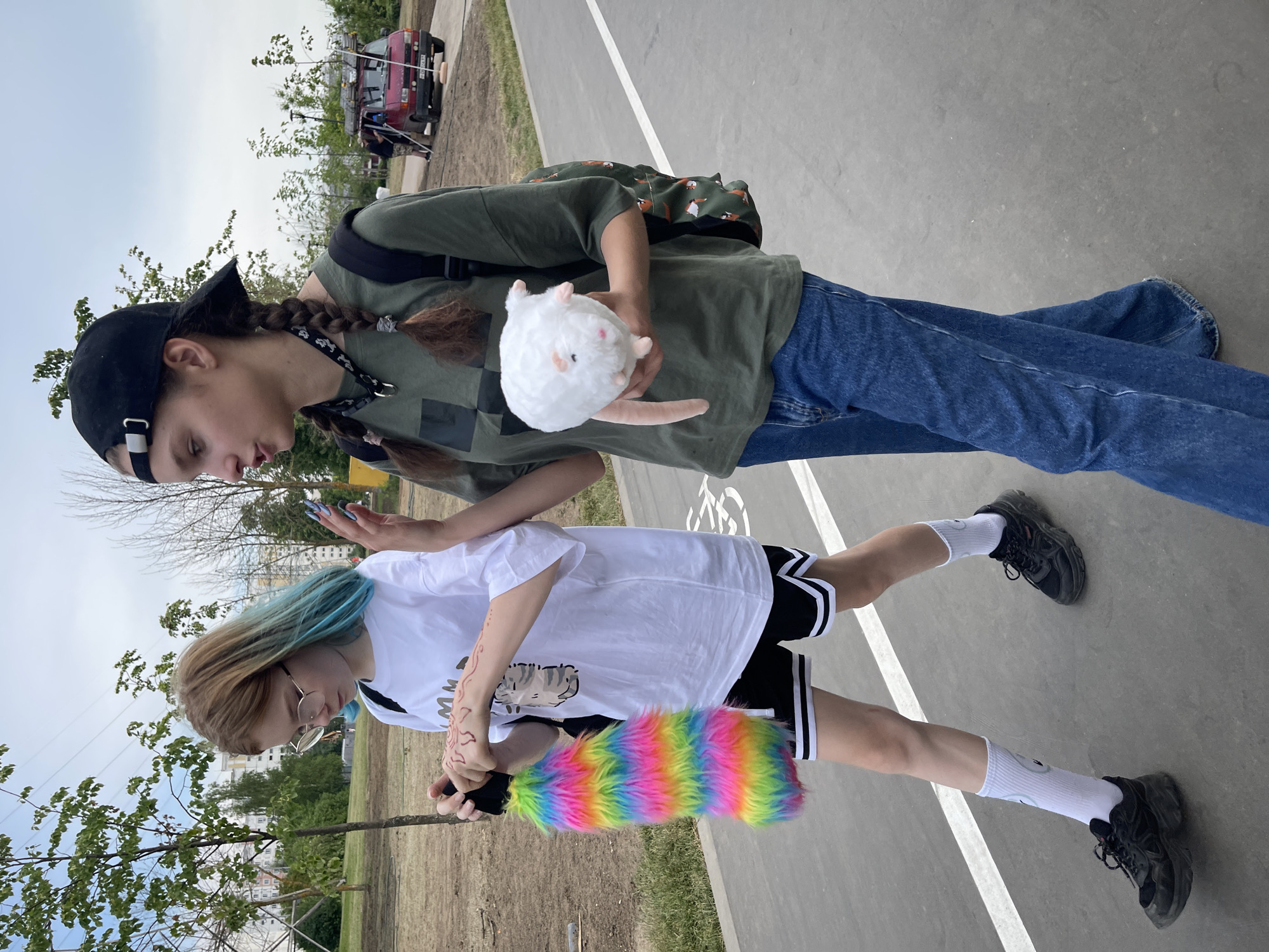 Two young women are walking on a paved path with white lane markings in a park-like urban setting. The woman on the left has blonde hair with blue streaks, wears glasses, a white t-shirt with a graphic print, black athletic shorts with white stripes, white socks, and black sneakers. She is holding a fluffy, rainbow-colored accessory. The woman on the right wears a dark green t-shirt, blue jeans, and a black baseball cap. She has dark hair braided into two pigtails and is carrying a plush white toy. Behind them, trees, buildings, and a red vehicle are visible. The sky is overcast. The setting appears to be a public park within the city of Moscow, Russia, possibly near residential buildings.