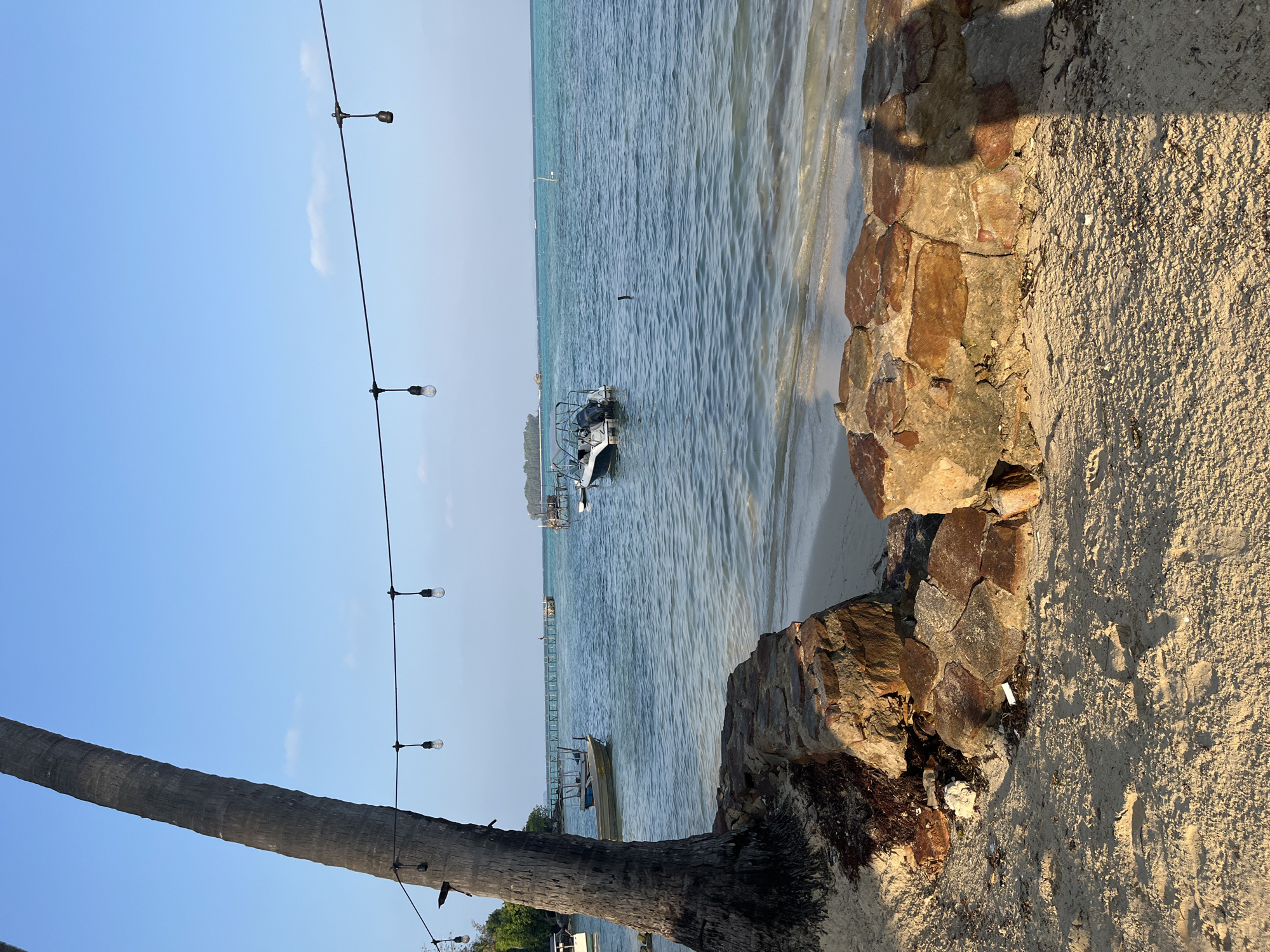 The image captures a serene beachfront scene at Legonipah, Indonesia. The foreground is dominated by a sandy beach with a low stone wall, partially covered by seaweed and debris. A large palm tree trunk is visible on the left, its bark textured and weathered. Above the beach and extending across the frame is a string of bare light bulbs, casting a warm glow.
The middle ground features the clear, calm turquoise water of the sea. Several boats are anchored or gently drifting: a small, light-colored boat with a canopy is moored closer to the shore, while a more modern speedboat with a white hull and roll cage sits further out. A wooden pier stretches into the water in the distance, with small structures visible at its end.
The background is a vast expanse of blue sky, with a few wispy clouds. The horizon is marked by distant islands, creating a sense of depth and remoteness. The lighting suggests it is either early morning or late afternoon, with the sun casting long shadows. The overall impression is one of tranquility and natural beauty, a typical coastal landscape in Indonesia.