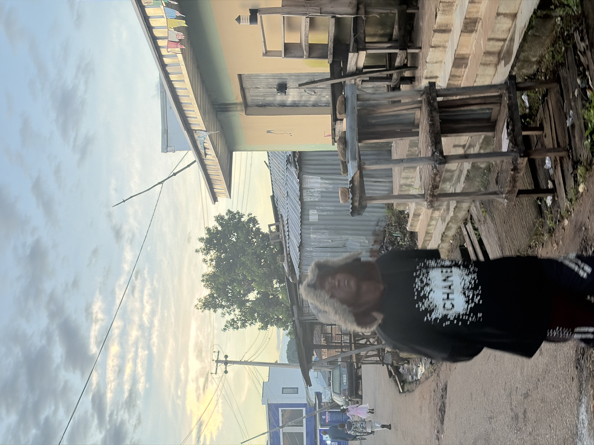 The image depicts a street scene in Jos, Nigeria, with a young person in the foreground wearing a black hoodie with a white "CHANEL" logo and a fur-lined hood. They are walking away from the camera. In the background, several corrugated metal structures and a light-colored building are visible, suggesting a commercial or residential area. A car is parked on the street, and other individuals are seen walking further down the road. The sky is cloudy with hints of sunset. A rustic wooden shelf unit is positioned prominently in the middle ground, adding to the utilitarian and informal atmosphere of the street.