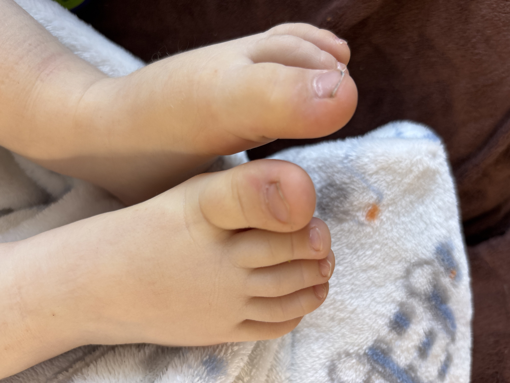 A close-up photograph shows two baby feet on a white blanket with light blue patterns. The background includes a brown surface. No details suggest the location is in Izhevsk, Russia.