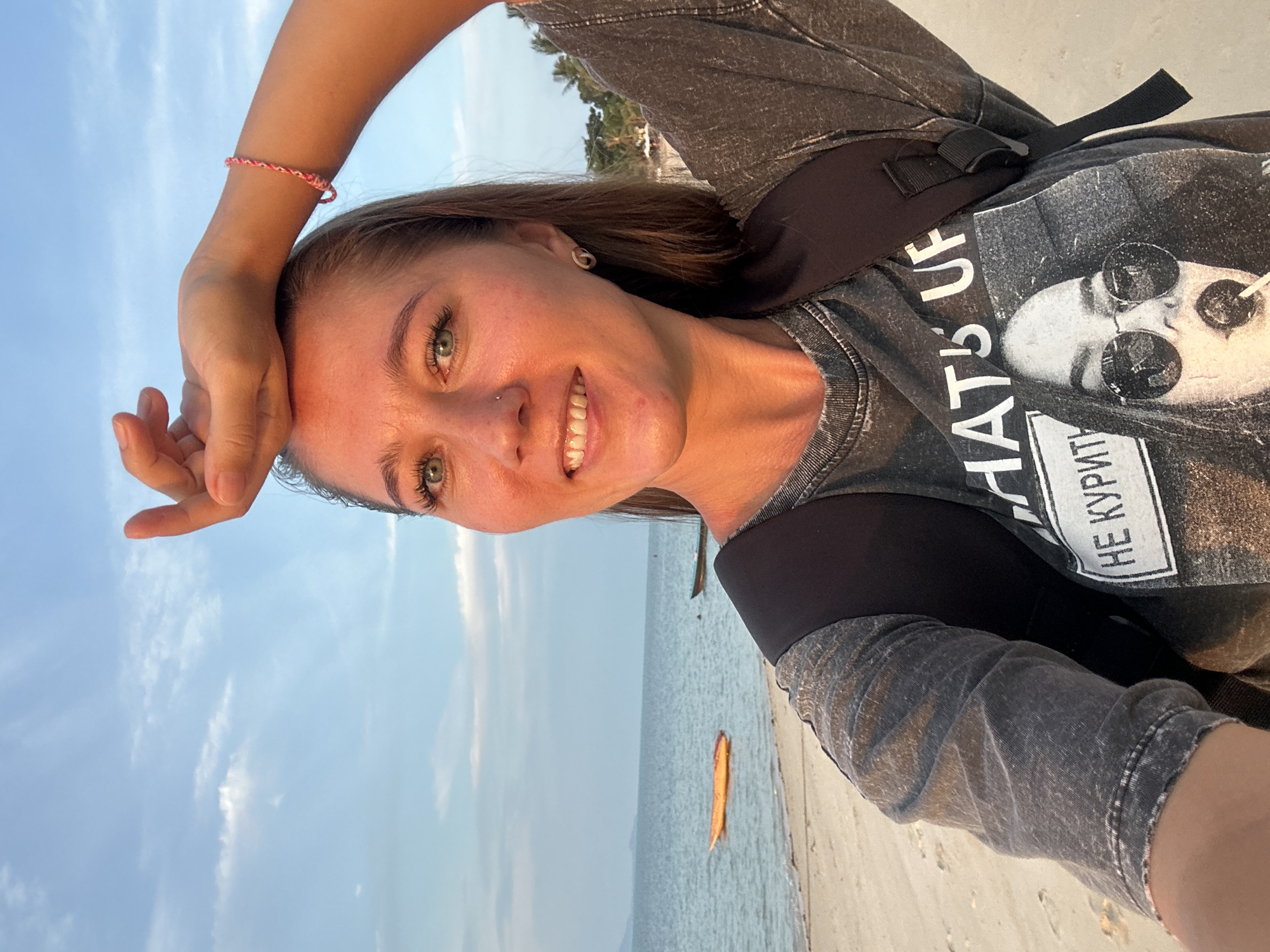 A young woman with light brown hair and blue eyes is taking a selfie on a beach in Ko Samui District, Thailand. She is smiling and has her right arm raised, with her hand on her forehead. She is wearing a dark gray t-shirt with a graphic print on it, and a black backpack. In the background, there is a sandy beach, the ocean, and a clear blue sky with some clouds. There are two boats in the water, one orange and one dark. The sun is setting, casting a warm glow on the scene.