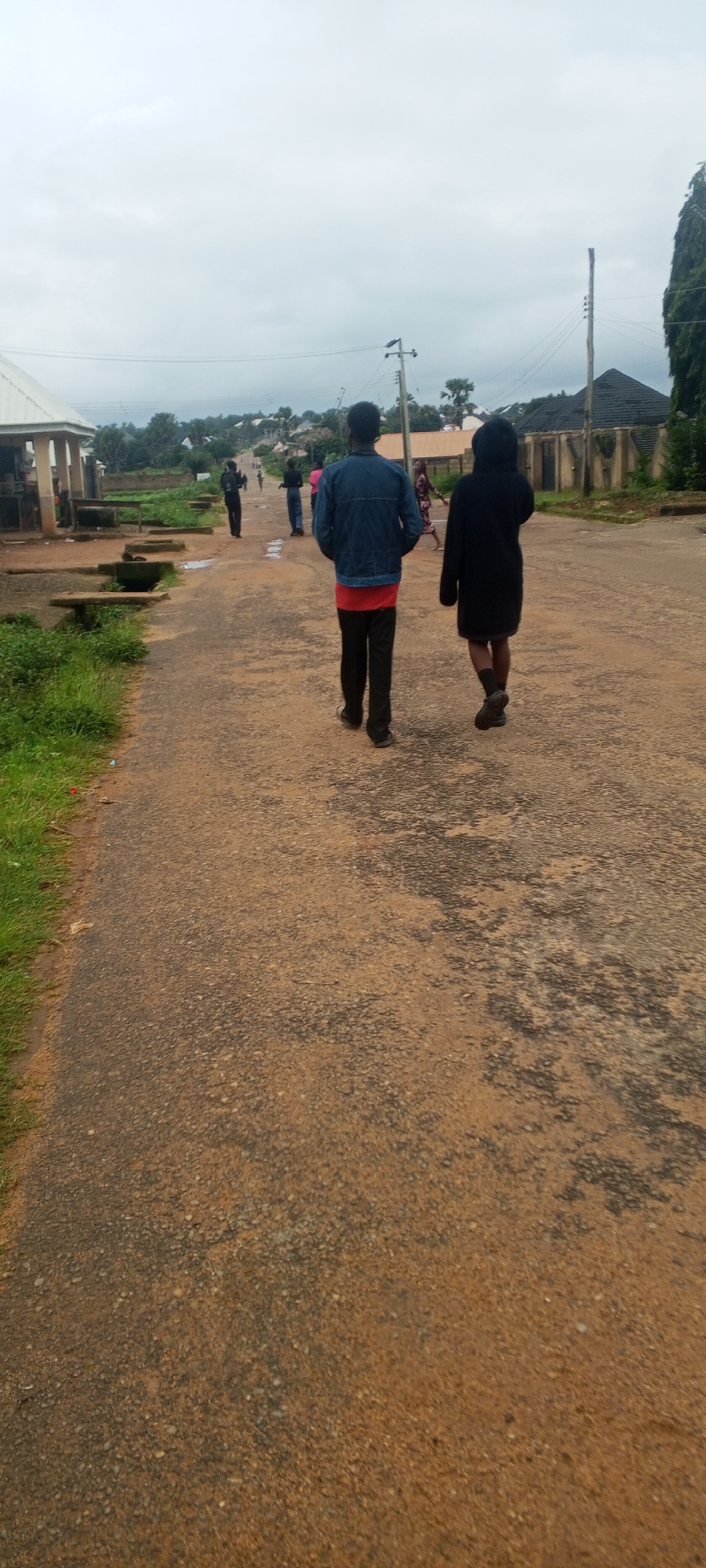 Two individuals walk away from the camera on a paved road in Bahit, Nigeria. The person in front is wearing a blue denim jacket over a red t-shirt and black pants. The person behind them is wearing a long black hooded coat. The road is surrounded by green grass and dirt on the left, and buildings and trees on the right. The sky is overcast and grey. In the background, several other people are visible walking in the distance, along with a utility pole and various structures.