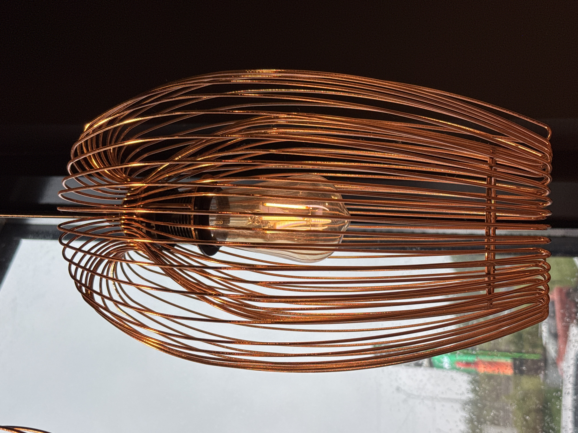 This is a close-up, eye-level shot of a decorative copper wire pendant light fixture, illuminated by a vintage Edison-style light bulb. The fixture is oval-shaped and composed of numerous vertical copper wires that curve inwards at the top and bottom, creating an open, cage-like structure. The light bulb, with its visible filament glowing warmly, is suspended at the center of the fixture.
The background is divided into two distinct areas. To the left, a window shows a blurred, overcast outdoor scene, possibly indicating rain or fog, with hints of green and orange signage and vehicles visible. The right side of the background is a solid, dark, out-of-focus area, likely representing an interior wall or curtain.
No people are present in the image, and no actions or interactions are depicted. The environment suggests an indoor setting with a view to the outside. There is no specific indication of the location being Medovoye, Russia, beyond the prompt.