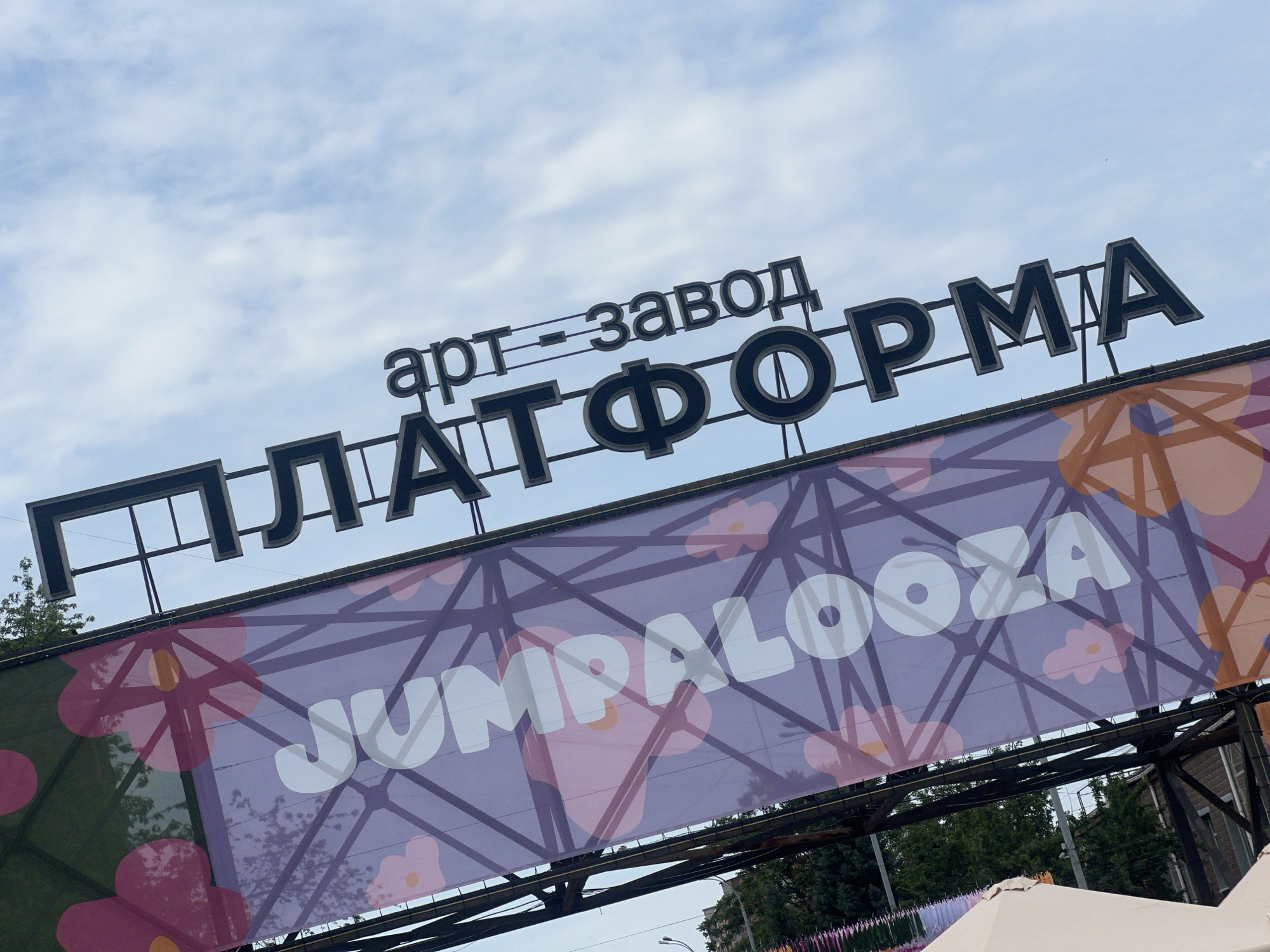The image displays an outdoor scene featuring prominent signage for an event venue in Kyiv, Ukraine. Against a light blue sky with scattered white clouds, large, three-dimensional dark grey Cyrillic letters are mounted on a structure. The top line reads "арт-завод" (Art Factory), and the line directly below it spells "ПЛАТФОРМА" (PLATFORMA). Below this permanent signage, a large, rectangular purple banner is stretched across a dark metal truss framework. The banner features a background design of interconnected geometric lines and stylized floral shapes in shades of pink and orange. Superimposed in the center of the banner, in large, rounded white capital letters, is the word "JUMPALOOZA". Green tree foliage is visible on the left and right sides of the image, behind and below the banner. In the lower left background, colorful hanging ribbons are discernible, while a partial view of a light-colored canopy or tent is present in the lower right foreground. No individuals are visible in the frame. The combined signage identifies the location as Art Factory Platforma (Арт-завод Платформа), a known cultural and event space in Kyiv.
