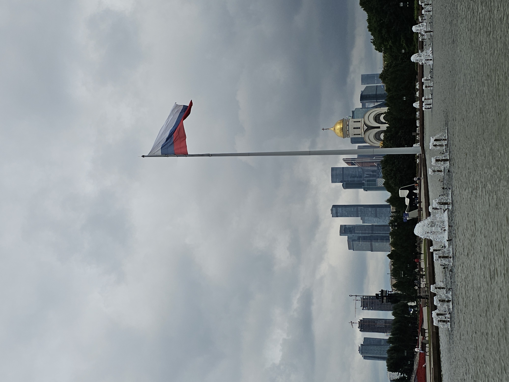 This image depicts a panoramic view of a park in Moscow, Russia, on a cloudy day. In the foreground, a large fountain is active, with numerous jets of water spraying upwards. Beyond the fountain, a paved walkway lined with benches and trees stretches across the middle ground. Several people are visible walking and sitting on the benches, engaged in leisurely activities. In the background, a cluster of modern skyscrapers forms the city's skyline, with a prominent golden-domed church situated among them. A tall flagpole flies the Russian flag, its tricolor stripes of white, blue, and red clearly visible against the overcast sky. The scene suggests a public gathering space within a major urban environment.