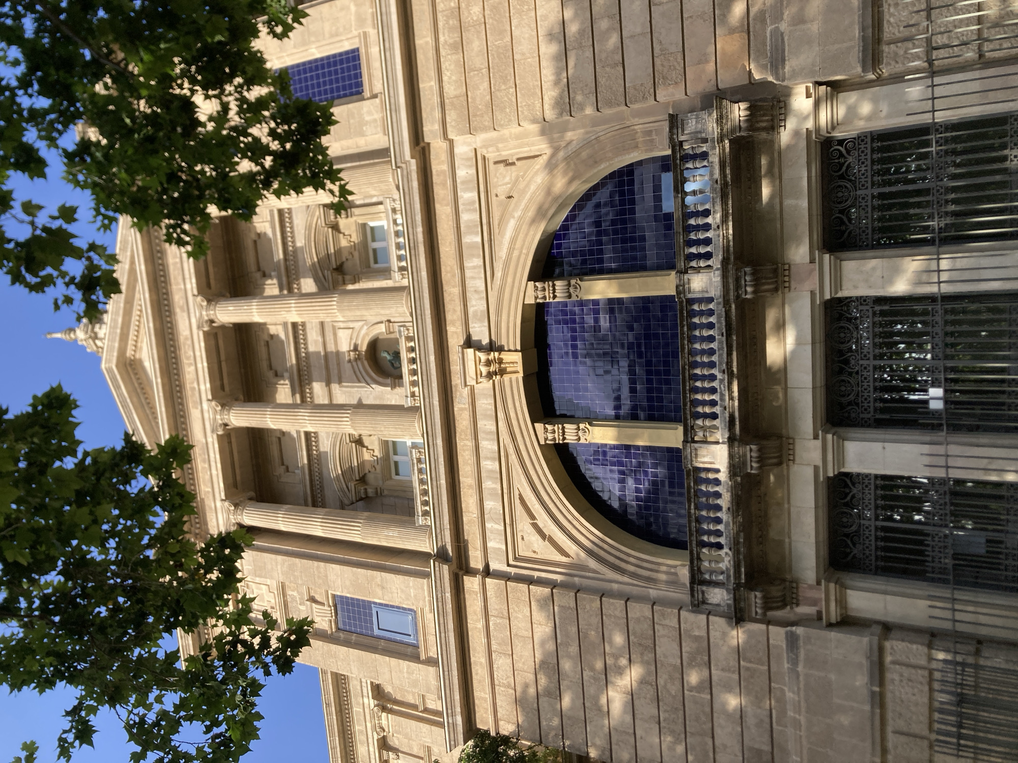 The image captures the exterior of the Palau de la Generalitat de Catalunya, a historical government building located in Barcelona, Spain. The perspective is low-angle, looking up at the facade. The building is constructed of light-colored stone blocks, with a neoclassical architectural style. Prominent features include large, fluted columns supporting an entablature, a balcony with ornate balustrades, and a large arched window filled with blue tiles that reflect the sky. Above the arched window, a smaller window is visible. The top of the building features a dome or cupola. Green tree branches with leaves hang into the frame from the top left and top right, partially obscuring the sky and the upper part of the building. The sky is a clear, bright blue. A black metal fence is visible in the foreground, partially obscuring decorative ironwork grates over doorways or ground-level windows. The lighting suggests it is daytime, with sunlight casting shadows on the stone facade. No people are visible in the image.