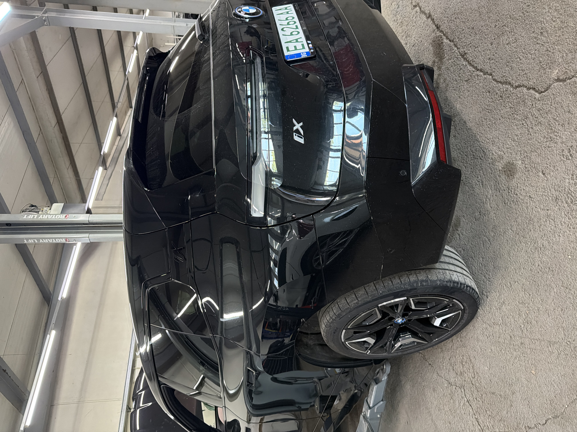 The image shows the rear passenger side of a black BMW iX parked in what appears to be a car repair shop or dealership. The car is on a lift, with its rear wheel exposed. The license plate is visible and reads "EA 6266 AA". The car's paint is highly reflective, showing the interior of the shop. The shop appears to have fluorescent lighting and a concrete floor. There is a "ROTARY LIFT" sign visible in the background. The location is identified as Burgas, Bulgaria.