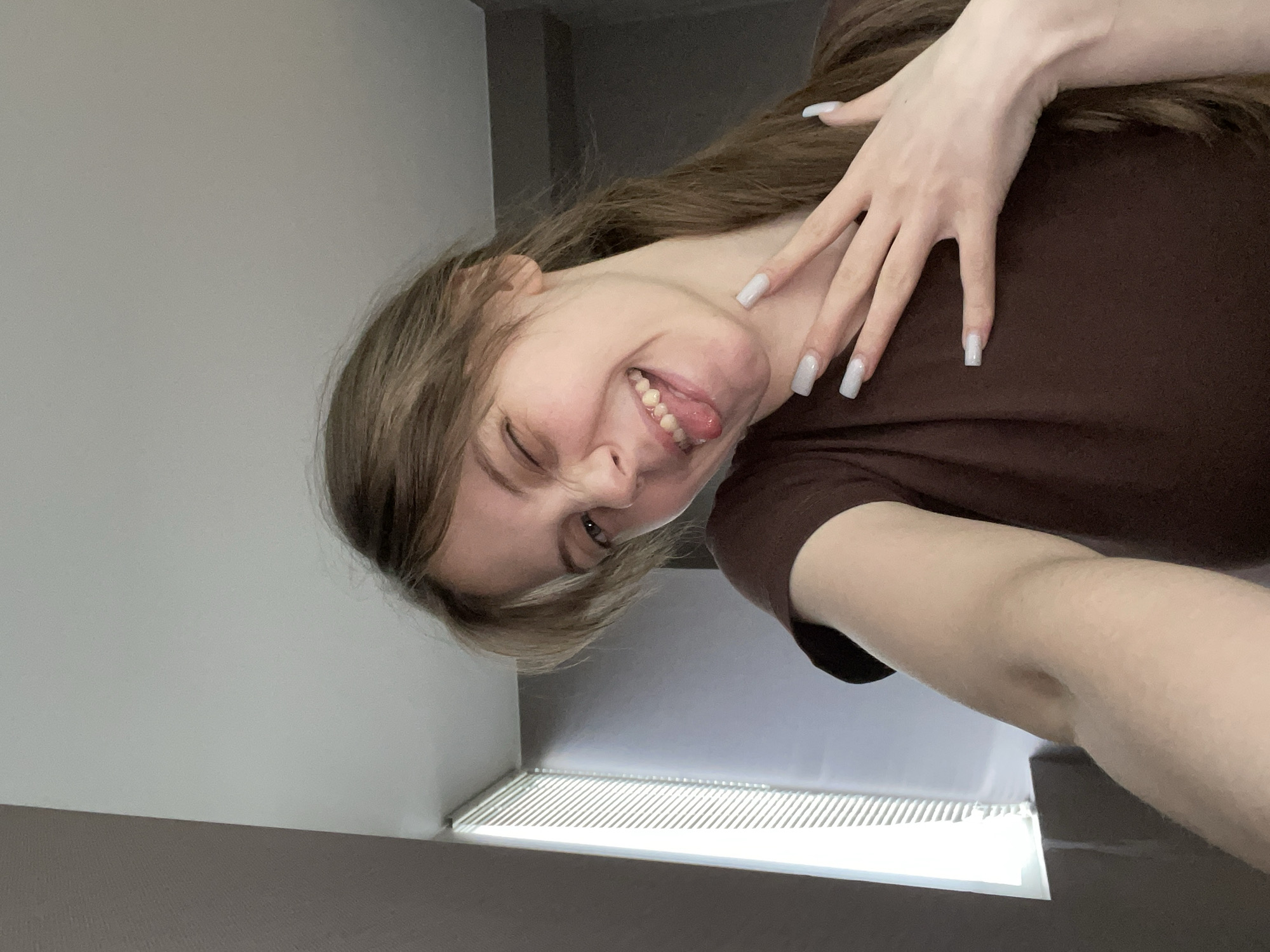 A woman is photographed in an upward-facing, close-up selfie. She has light skin, brown hair, and is sticking her tongue out while winking one eye. Her right hand, with long, rectangular nails, is raised near her neck. She is wearing a brown shirt. The background consists of a gray wall and a light fixture with horizontal slats. The location is identified only as the city of Ufa, Russia. There are no other people or objects clearly visible in the image.