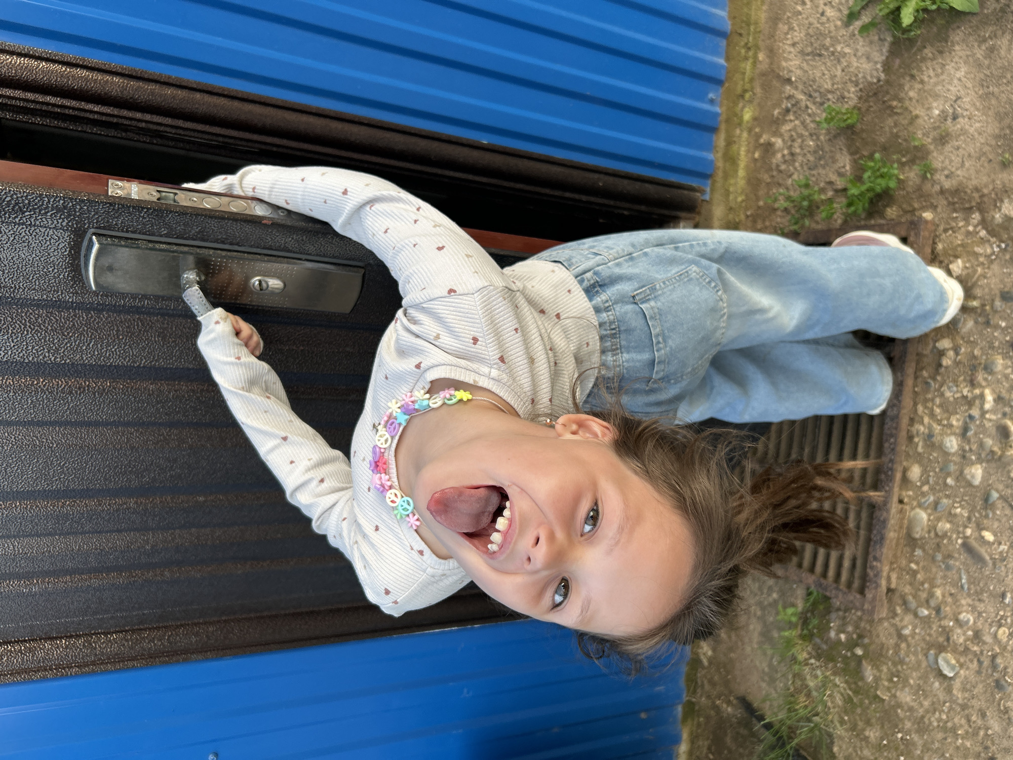A young girl, with brown hair tied in a ponytail and a multi-colored beaded necklace, sticks out her tongue. She is wearing a long-sleeved shirt with small hearts and light blue denim jeans. She is reaching out with her right hand towards a door handle. The door appears to be a dark metal with a textured surface. Behind the girl is a blue corrugated metal wall and a patch of gravel and grass. The scene suggests an outdoor setting. The lighting appears to be natural, indicating it is daytime, possibly sunny. The girl's expression is playful and energetic. The crops show details of the door lock mechanism and the texture of the blue wall. No visible text is discernible in the image or crops. The location context is Фоки, Russia.