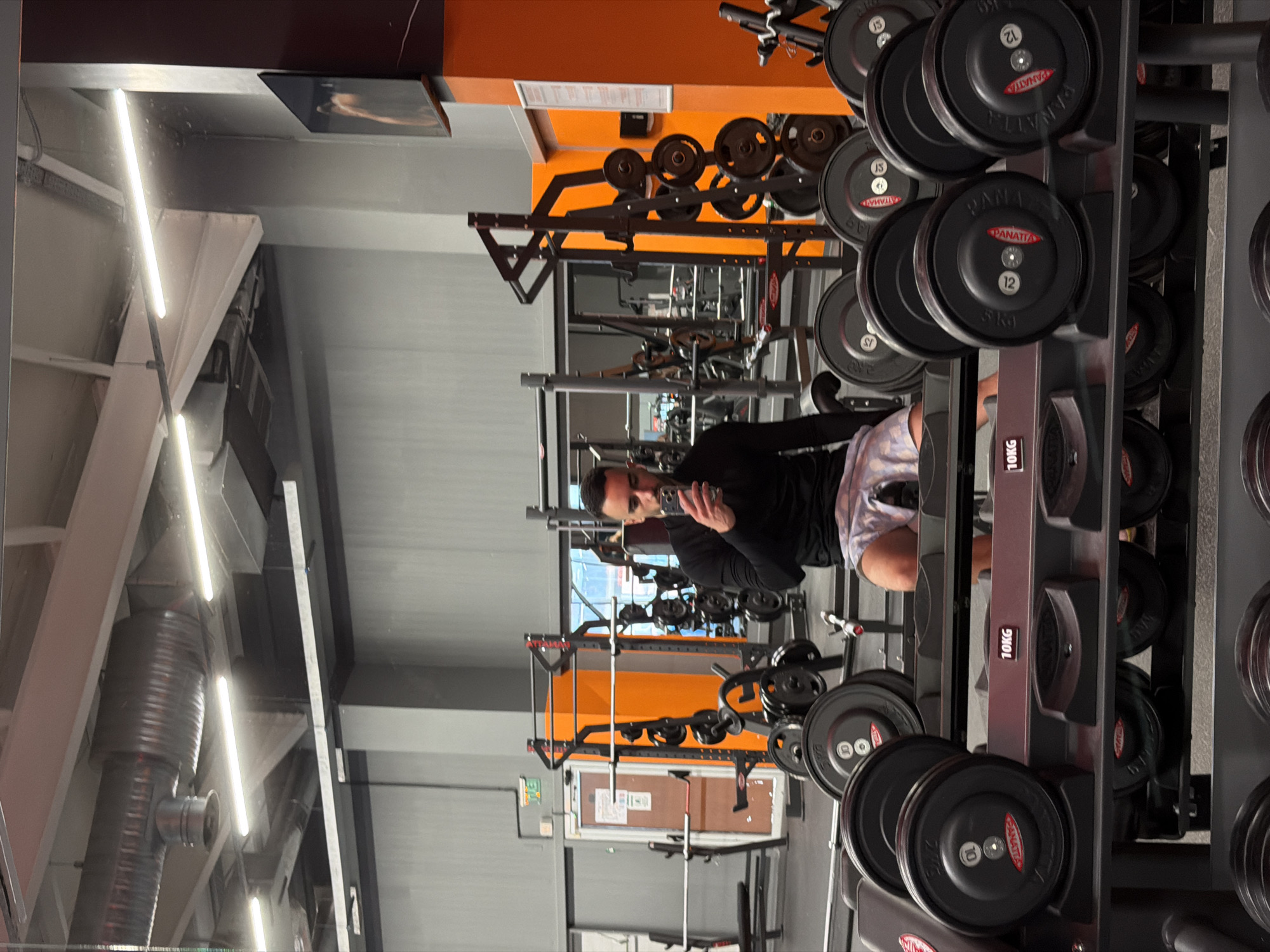 A man is taking a selfie in a gym. The gym has weight racks filled with black and grey weight plates. In the background are other gym equipment such as squat racks and weight machines. The ceiling has exposed pipes and industrial lighting. The gym has orange and grey walls. There is a door in the background. The man is wearing a black long-sleeved shirt and patterned shorts. The gym is located in the city of Plovdiv, Bulgaria.