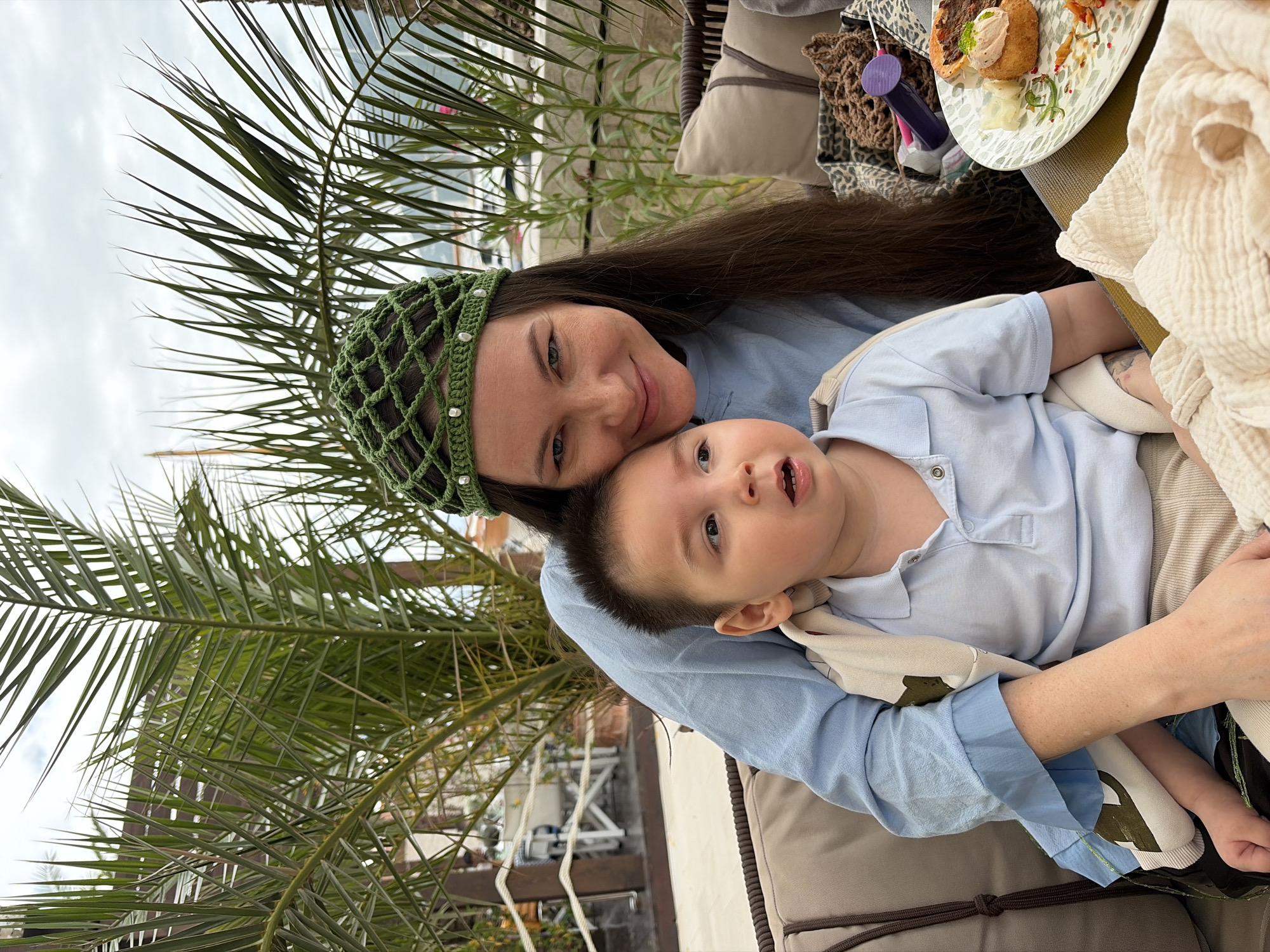 A woman wearing a green crocheted headpiece sits outdoors, holding a young child in her lap. Both are dressed in light blue clothing. A plate of food sits on a table to their right, alongside a collection of personal items. Palm leaves are visible in the background. The location is Đenovići, Montenegro, as it is specified in the task, but does not appear in the image.