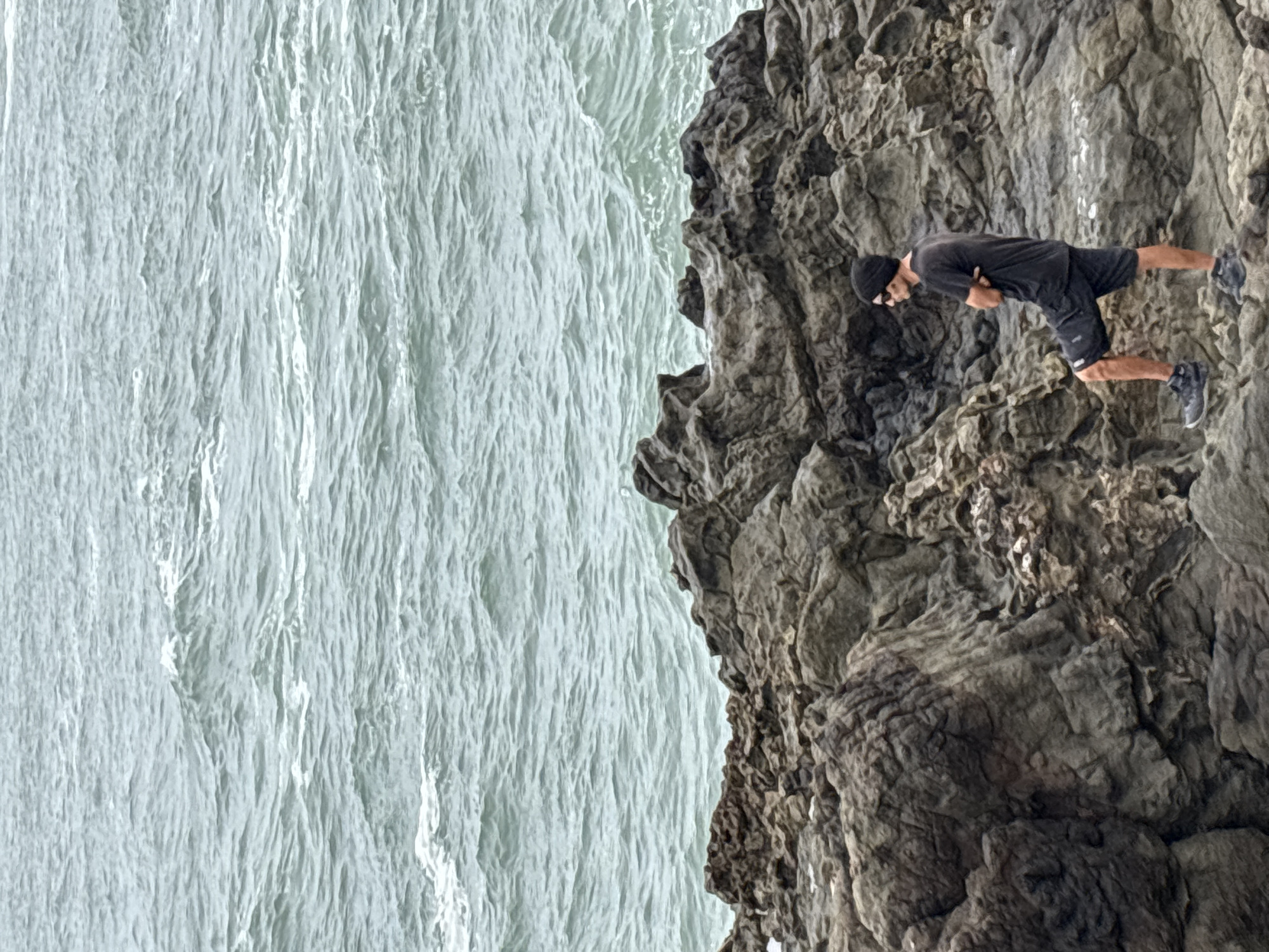 A man wearing a black t-shirt, black shorts, and a black beanie, with sunglasses on his face, stands on a rocky outcrop by the sea. He has his arms crossed and appears to be looking out at the ocean. The water is a light, greenish-gray color, with visible ripples and small waves. The rocks are dark gray and rugged. It is likely that the image was taken in Arica, Chile, near the coastline.