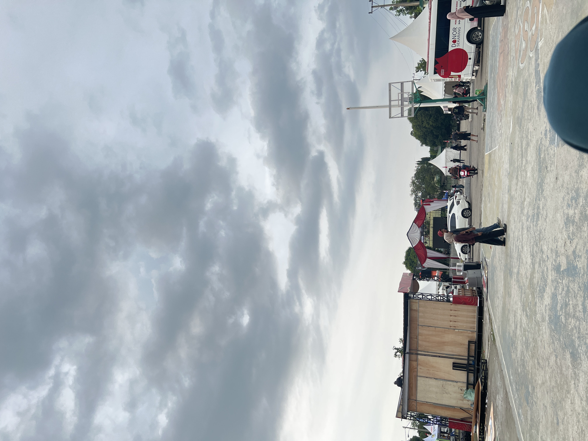This is a wide-angle, eye-level outdoor photograph taken on a cloudy day in the city of Lhokseumawe, Indonesia. The foreground shows a basketball court with faded painted lines and some circular markings. In the mid-ground, a wooden stage structure is being assembled or dismantled, with scaffolding and various materials around it. Two individuals, a man and a woman, are walking across the court in the mid-ground, both wearing casual clothing. A white bus with red accents and the text "DOHOR SEKARANG" and "PMI" is parked on the right side of the image, suggesting a blood donation event. Several white tents are set up behind the bus, and other vehicles, including cars and a motorcycle, are visible. In the background, trees and utility poles can be seen. The sky is filled with heavy, gray clouds, indicating overcast conditions. The overall scene depicts an outdoor event setup or breakdown in what appears to be a public space or sports complex.