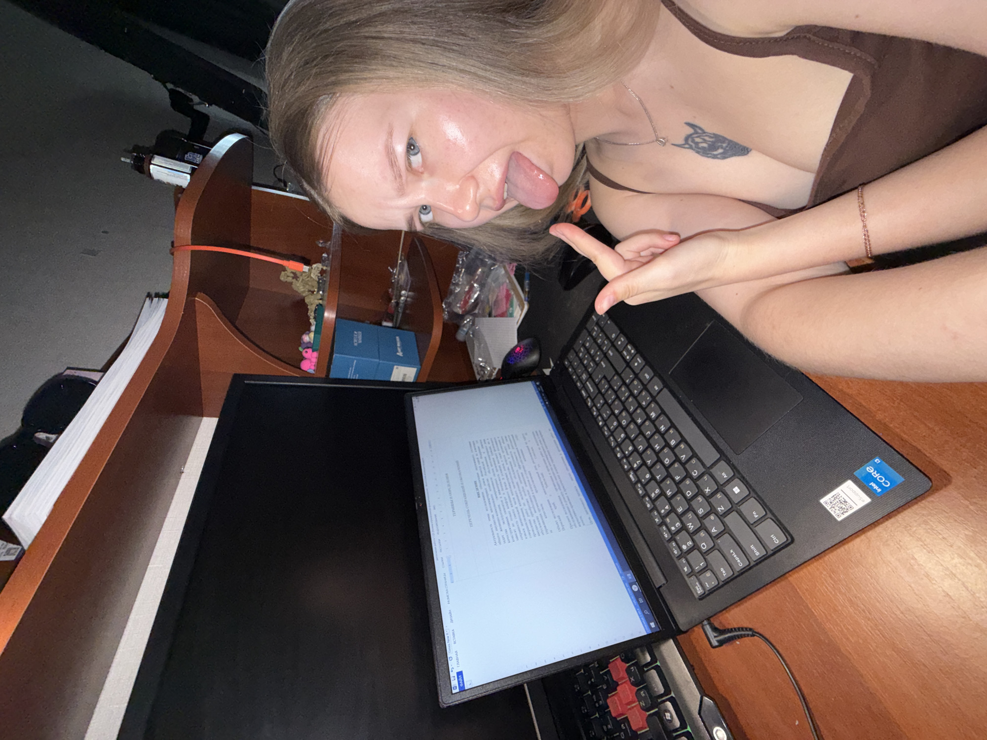 Here's a forensic description of the image:
**People:** A young adult female is present. She has blonde hair and fair skin. She is sticking out her tongue and making a gesture with her hand. She is wearing a brown top and a necklace. A tattoo is visible on her chest.
**Subjects:** A desk or table is the primary surface.
**Objects:** A laptop is open on the desk, displaying a document in a word processor. A second monitor is present and turned on but shows a blank screen. A gaming keyboard and mouse are below the monitor. A book shelf and other objects are in the background.
**Actions:** The female is interacting with the environment. Her expression and gesture suggest a playful or humorous pose.
**Interactions:** The person interacts with technology.
**Environment:** The setting appears to be indoors, possibly a home office or study area. The lighting is dim.
**Location:** The location cannot be determined to be Chaikovsky, Russia from the image.