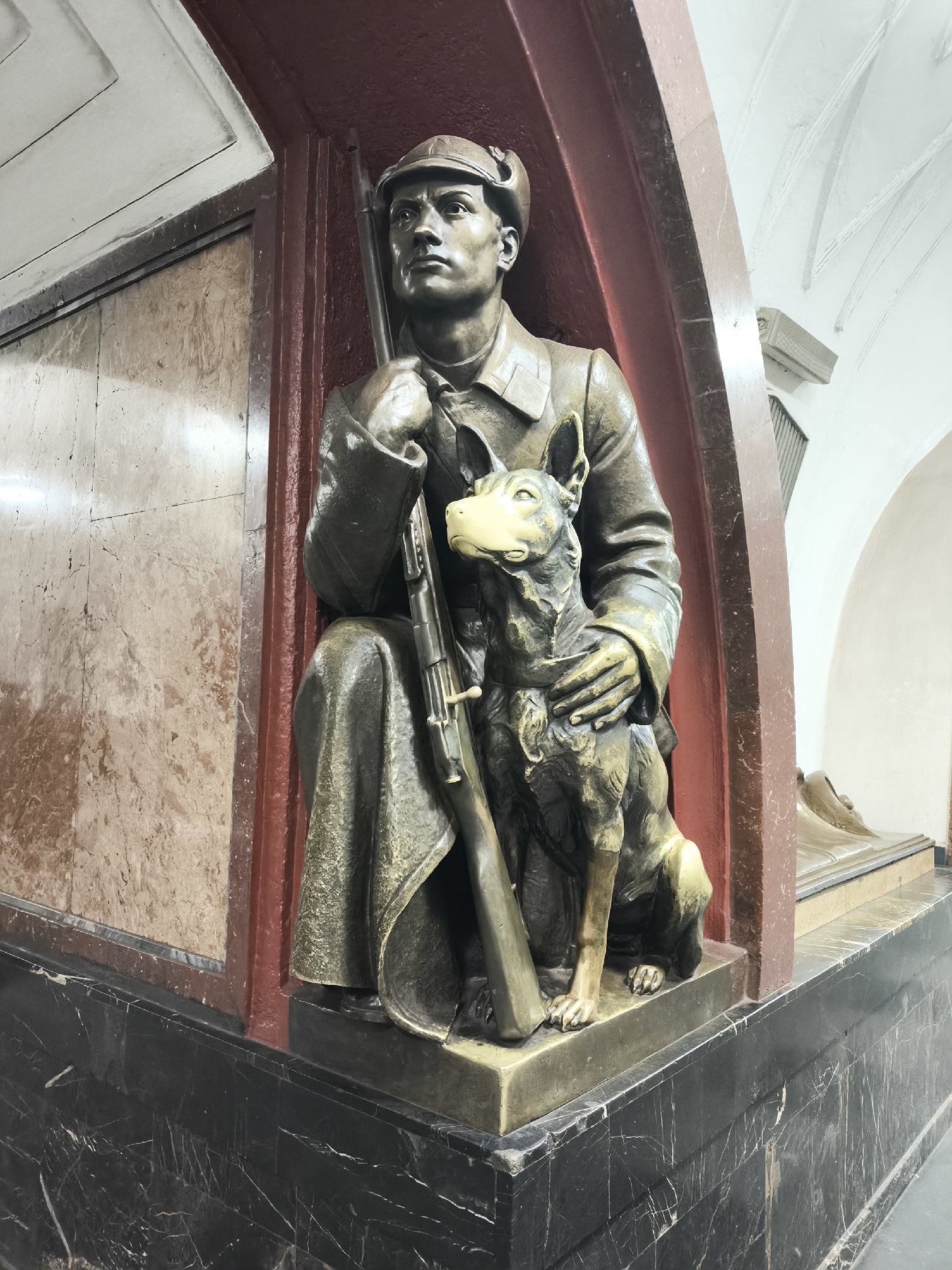 A bronze sculpture of a man and a dog is prominently displayed within an ornate indoor architectural setting. The sculpture occupies a corner, set against a dark reddish-brown recessed panel.
The man, depicted in a squatting or kneeling posture, wears a cap and a uniform-like jacket and trousers. He holds a long rifle vertically, resting it against his left leg while both hands grasp it. His face is chiseled, with a serious and contemplative expression, gazing upwards and slightly to the right.
Beside him sits a medium-sized dog, possibly a German Shepherd, looking upwards with an alert and attentive gaze. Its muzzle appears lighter in color, suggesting frequent touching or a different material/patina. The man's right arm is around the dog, indicating a close bond.
The sculpture rests on a dark, possibly black, marble base featuring white veining. To the left of the sculpture, the wall is clad in a lighter, beige or pinkish-brown marble with intricate natural patterns and veining. To the right, beyond the reddish-brown panel, white arched structures and ceiling sections are visible, implying a larger, grand hall or passageway. The architectural style suggests a monumental public space, such as a metro station.
The lighting appears artificial, highlighting the bronze details and marble textures. No visible text is present in the image.