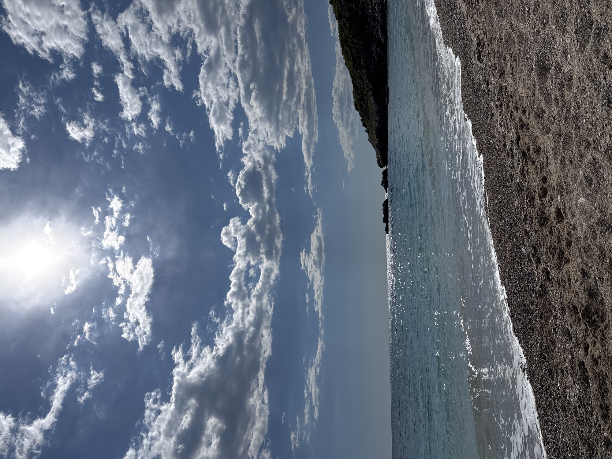A horizontal outdoor shot shows a beach in Rađenovići, Montenegro, on a sunny day. The sky, dominated by a bright sun, is filled with white, fluffy clouds against a blue background. The water, a clear, turquoise color, laps gently at the dark sand of the beach. A rocky cliff covered in green vegetation is visible in the distance. There are no people in the scene.