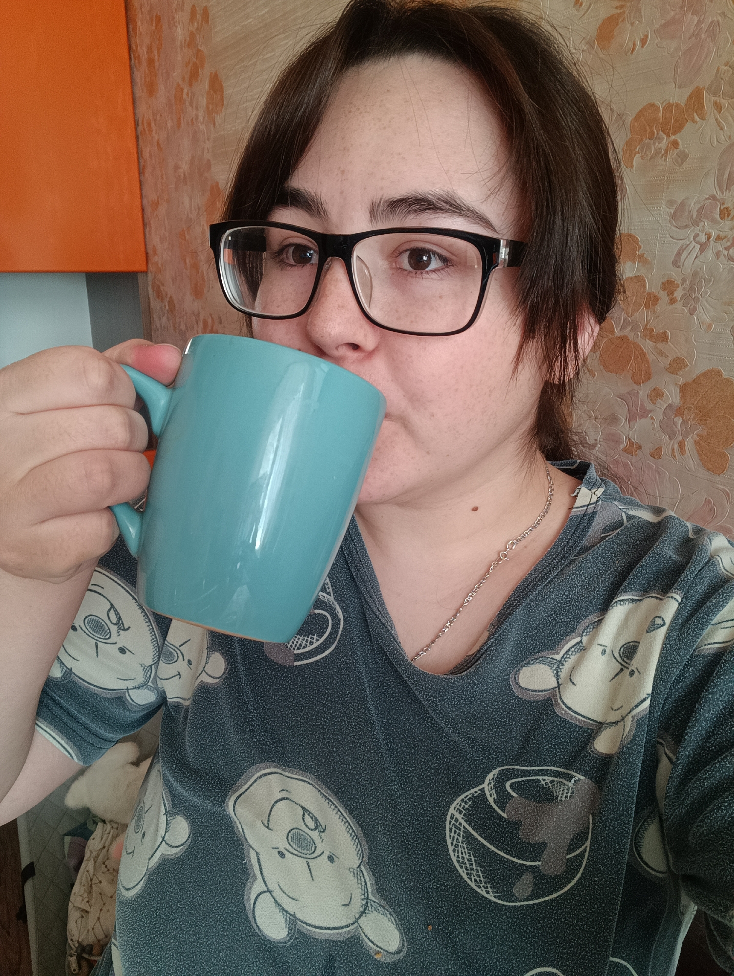 A woman with dark brown hair and black-framed glasses is depicted indoors, taking a sip from a large, bright teal-colored mug. She holds the mug with both hands, its rim touching her lower lip. Her expression is neutral and focused on the action. She has fair skin with visible freckles and wears a casual short-sleeved top patterned with light-colored, stylized animal faces and other symbols, possibly resembling cartoon bears or similar figures. A thin silver chain necklace is visible around her neck.
The background features an orange, rectangular cabinet or piece of furniture to the left. To the right and behind her, there is a wall covered with light brown and beige floral or vine-patterned wallpaper. A hint of a white, furry animal (likely a cat or small dog) can be seen behind her left arm, resting on a surface. The lighting suggests it is daytime. The overall scene conveys a casual, relaxed atmosphere in what appears to be a home in Noviy, Russia. There is no visible text in the image.