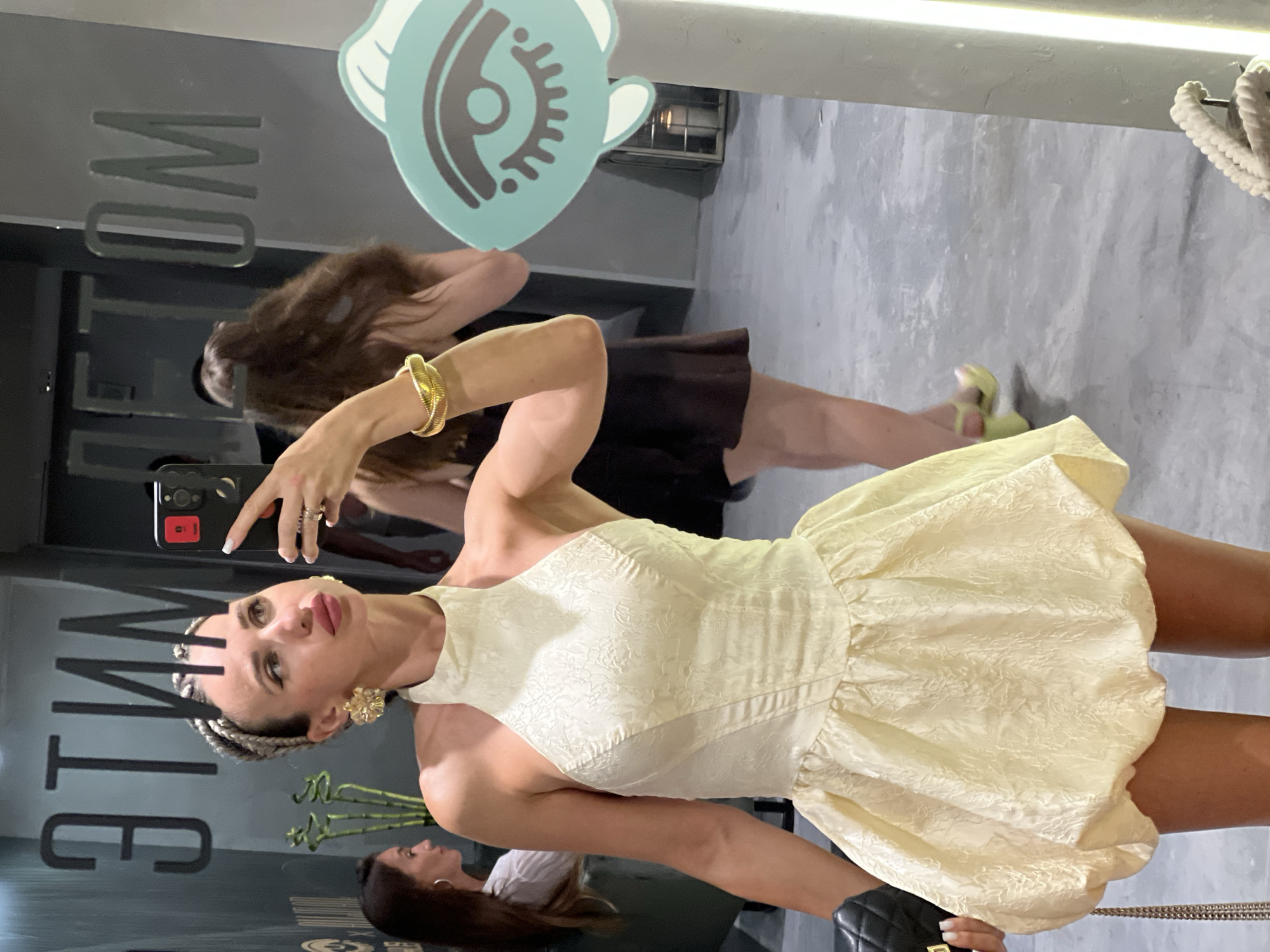 A woman with braided hair and large, floral gold earrings is taking a mirror selfie in what appears to be a modern establishment. She is wearing a cream-colored halter dress with a fitted bodice and a voluminous, textured bubble skirt. She holds a black quilted purse with a chain strap in her left hand and a smartphone in her right. In the background, reflected in the mirror, are other individuals and signage. To the left, a woman in a white shirt is visible, and further back, a woman with long, dark hair is standing. The mirrored surface displays text that appears to read "TIM ETOM" backward, possibly indicating a location or brand. A teal decal of an eye is affixed to a pillar to the right of the frame. The floor is a polished concrete surface. The scene suggests an indoor setting, potentially a bar, restaurant, or boutique, located in Sochi, Russia, given the presence of the name "Sochi" on the mirror.