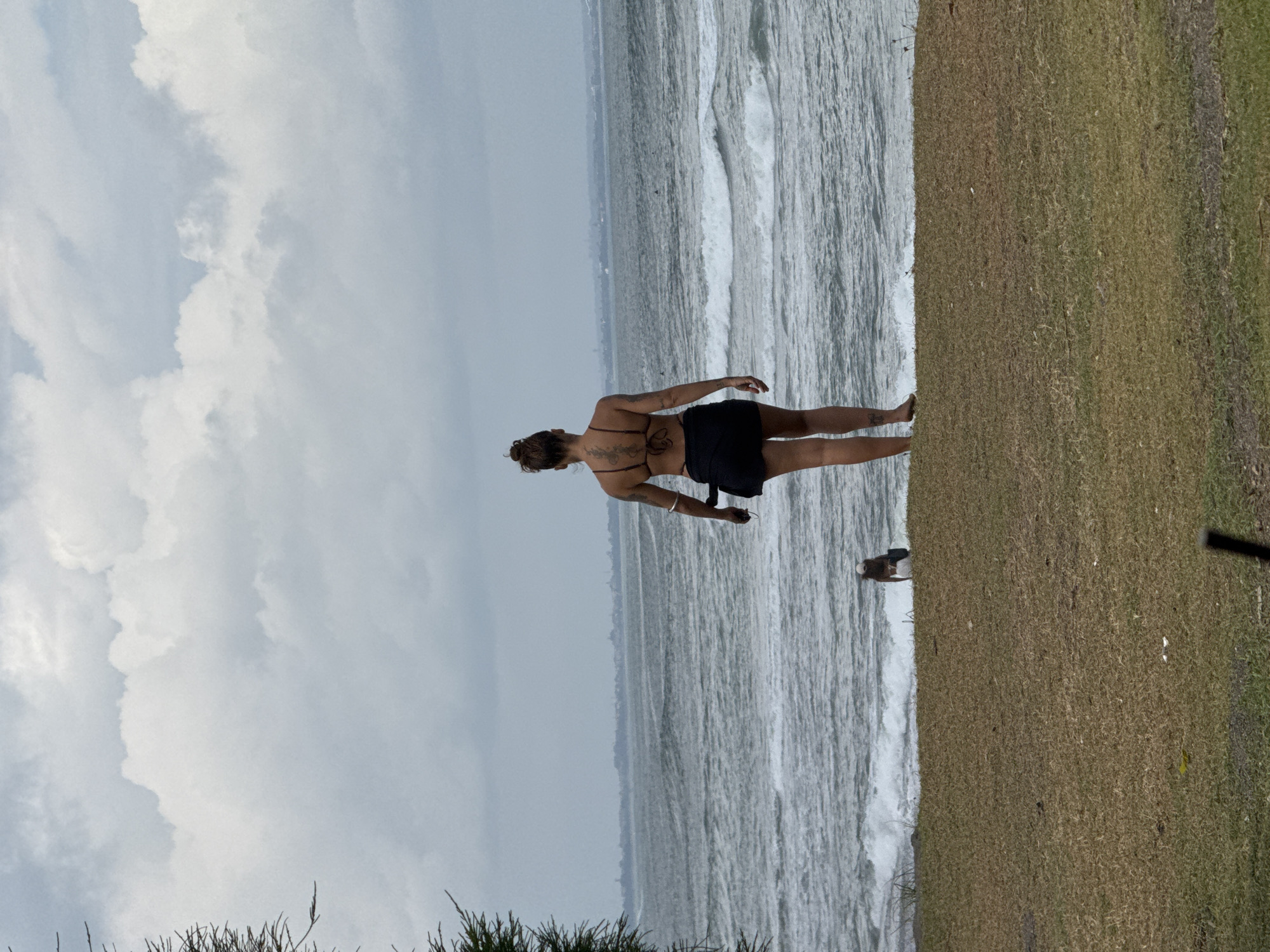 In Mengwi, Indonesia, on an overcast day, a woman with tattoos on her back stands facing a choppy ocean with grey skies. She is wearing a dark bikini top and a dark skirt. To her left, a person is crouched down near the water's edge. The foreground shows a grassy embankment.