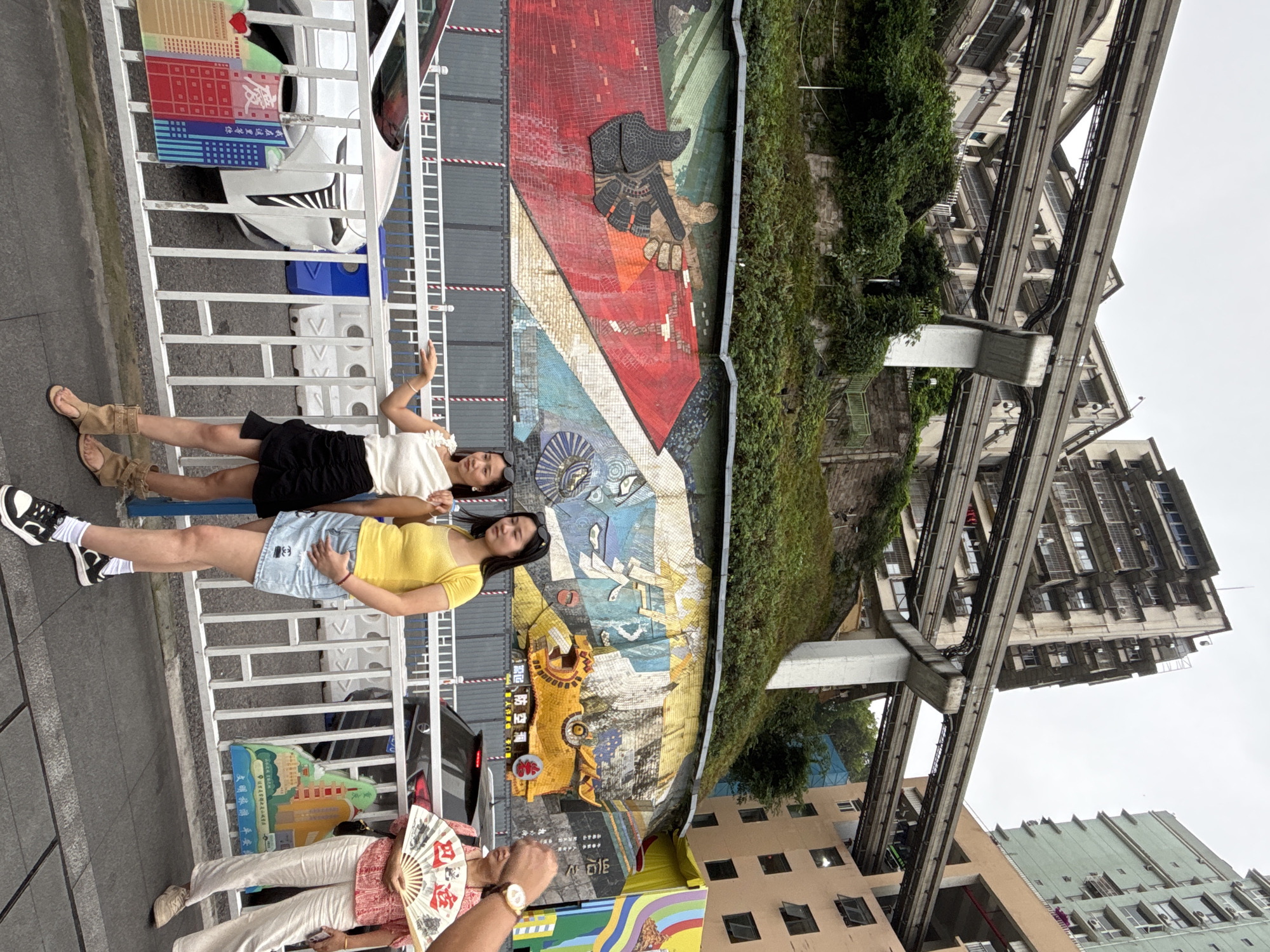 The image shows a street scene in Yuzhong District, Chongqing, China. Two young women are posing for a photo in front of a large mosaic mural that depicts stylized figures and patterns in red, blue, yellow, and other colors. They are standing behind a white metal railing. One woman is wearing a yellow top and denim shorts, and the other is wearing a white top and a black skirt. An older person is holding a folding fan with Chinese characters on it, seemingly taking the photo. A series of elevated train tracks runs along the buildings, which are mid-rise apartments with air conditioning units. The weather is overcast.