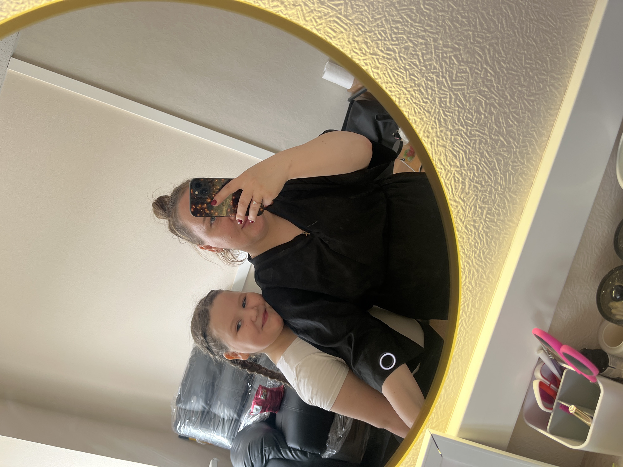 A mirrored selfie depicts a woman and a child in what appears to be a domestic or commercial setting, possibly within the city of Chaikovsky, Russia. The woman, holding a smartphone with a patterned case, is taking the photo. She has her hair in a bun and is wearing a black robe or garment. The child, positioned closer to the bottom of the frame, smiles at the camera. The child has braided hair and wears a white t-shirt. In the background, a black leather chair wrapped in plastic is visible, suggesting possible furniture delivery or renovation. A white storage container with various beauty or hair styling tools, including pink scissors, is also visible in the mirror's reflection. The mirror itself has a gold frame. The wall behind the mirror is covered with textured wallpaper.