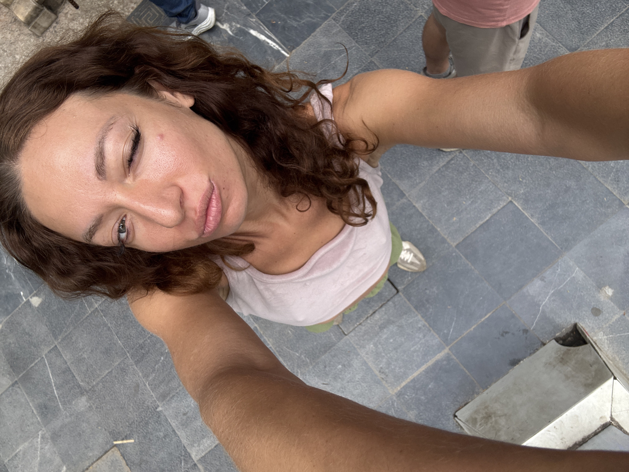 A woman with curly brown hair is taking a selfie outdoors in Hà Giang 1 Ward, Vietnam. She's wearing a light pink tank top and green shorts. Her left eye is closed in a wink. The ground is covered in gray and blue square tiles. Two other people are partially visible: one wearing gray shorts and the other wearing pink shorts.
