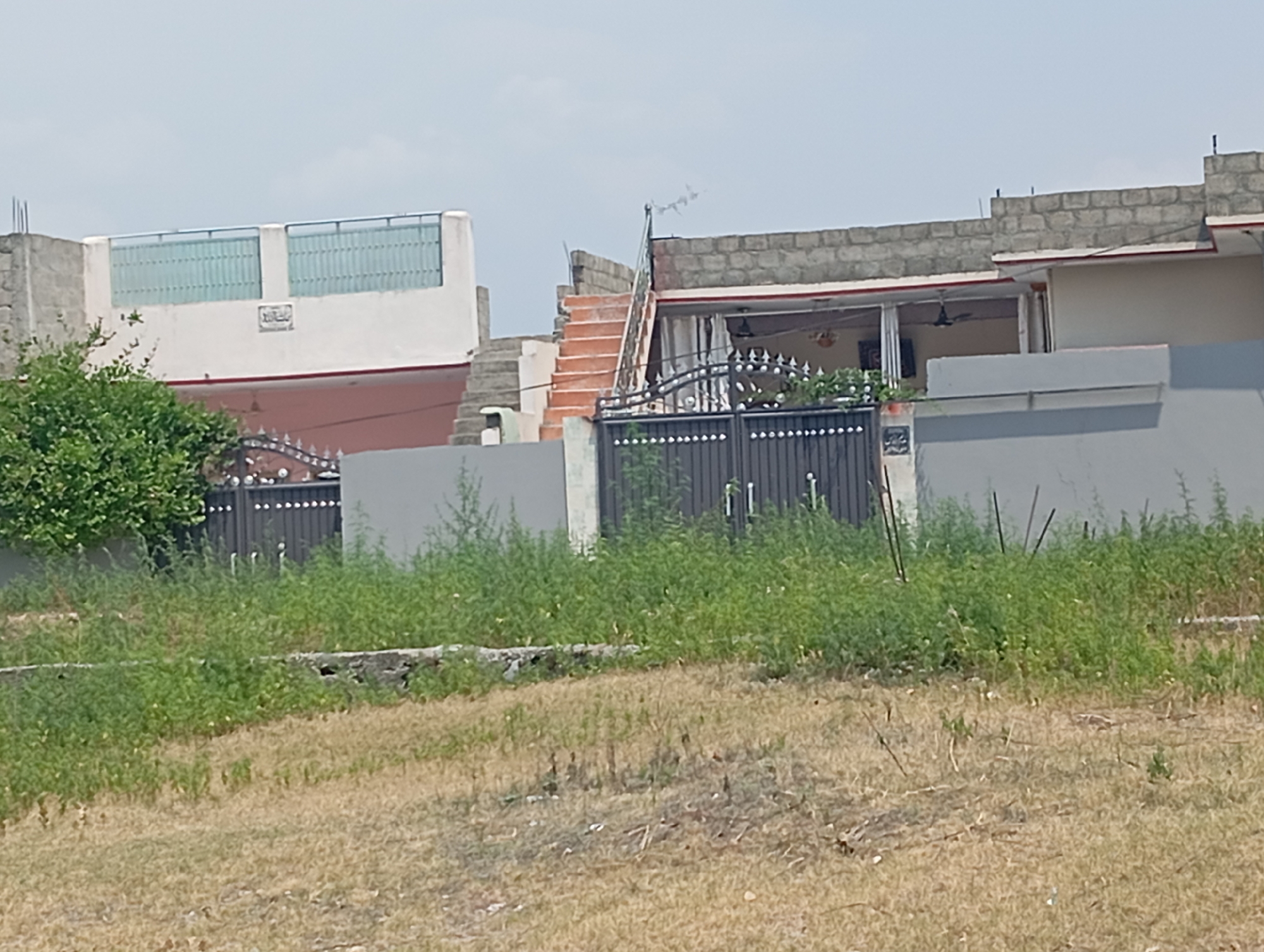 In Hakale, Pakistan, an outdoor scene depicts a residential property with a gray metal double gate in the foreground, flanked by tall green vegetation. Beyond the gate, a portion of a building is visible, featuring a white upper facade with two large windows and an inscription. Adjacent to this, a section of a brick building with a red trim and an open doorway shows a ceiling fan. A set of orange stairs with a metal railing leads up to another part of the property. The background is dominated by a hazy sky. The ground in front of the gate is covered with dry grass and some scattered debris.