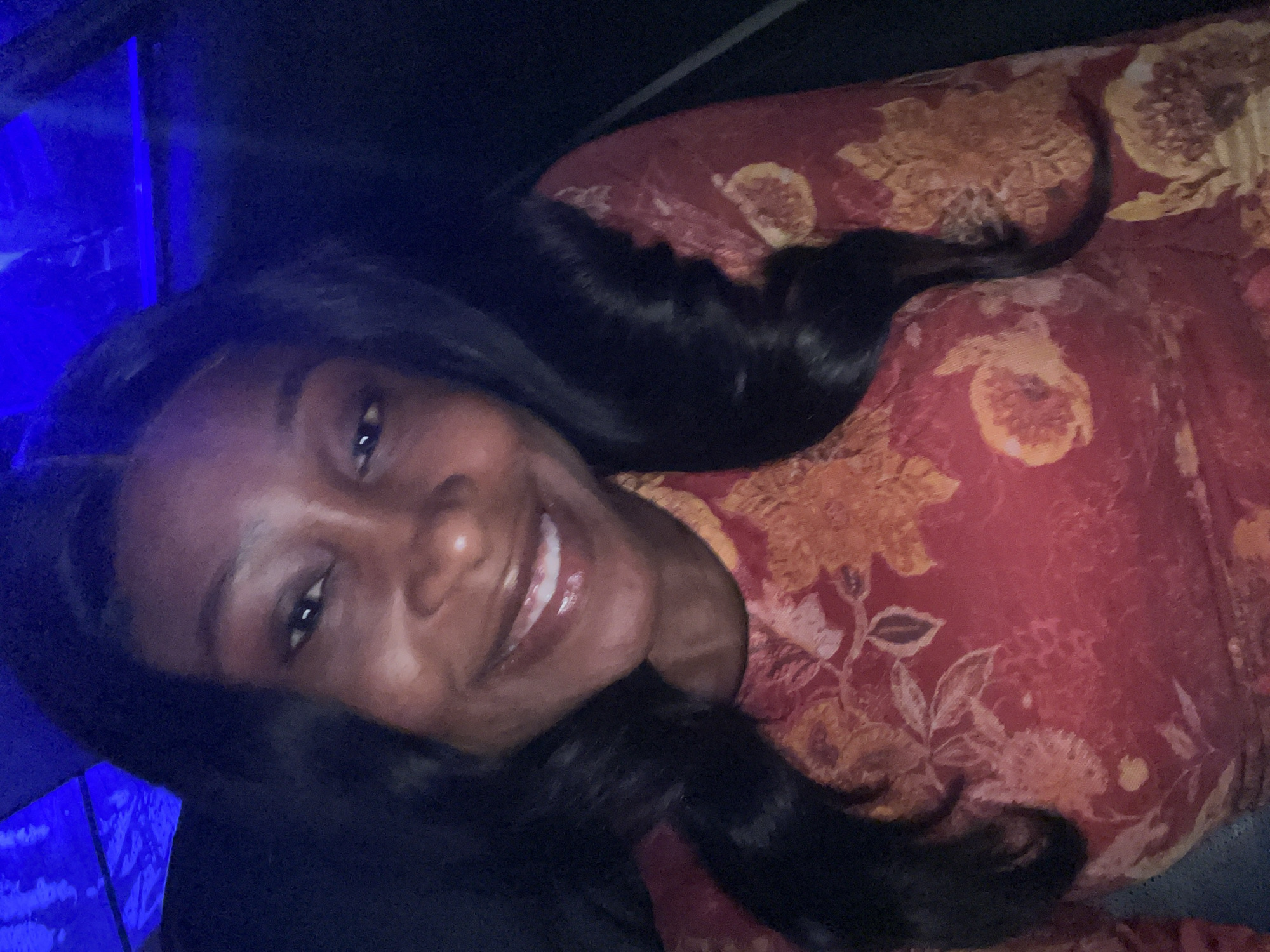 A close-up, waist-up photograph captures a Black woman with long, dark, wavy hair, smiling broadly. She is wearing a red long-sleeved top with a floral pattern in shades of orange and yellow. The background is dark with an overhead blue light. The image appears to be a selfie taken indoors, possibly in a vehicle or a dimly lit room. There is no indication of the location being Abuja, Nigeria.