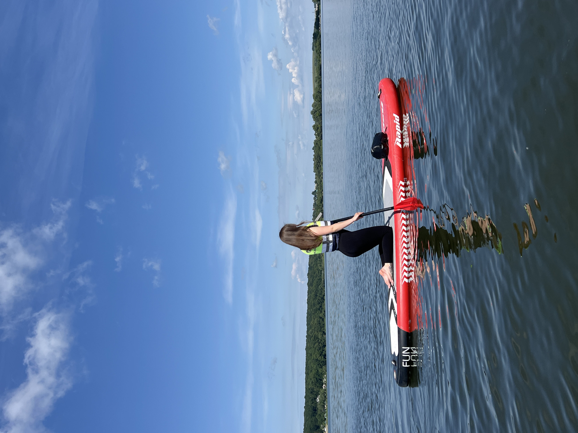 A woman is kneeling on a red paddleboard in the middle of a calm body of water. She is wearing a yellow and black life vest over a black top and black leggings. Her hands are on a red paddle, which is in the water next to her. The paddleboard has "FUN" and "RAPID" written on it in white lettering. The water reflects the blue sky with white clouds and the green tree line in the distance. The location is not Ternopil, Ukraine.